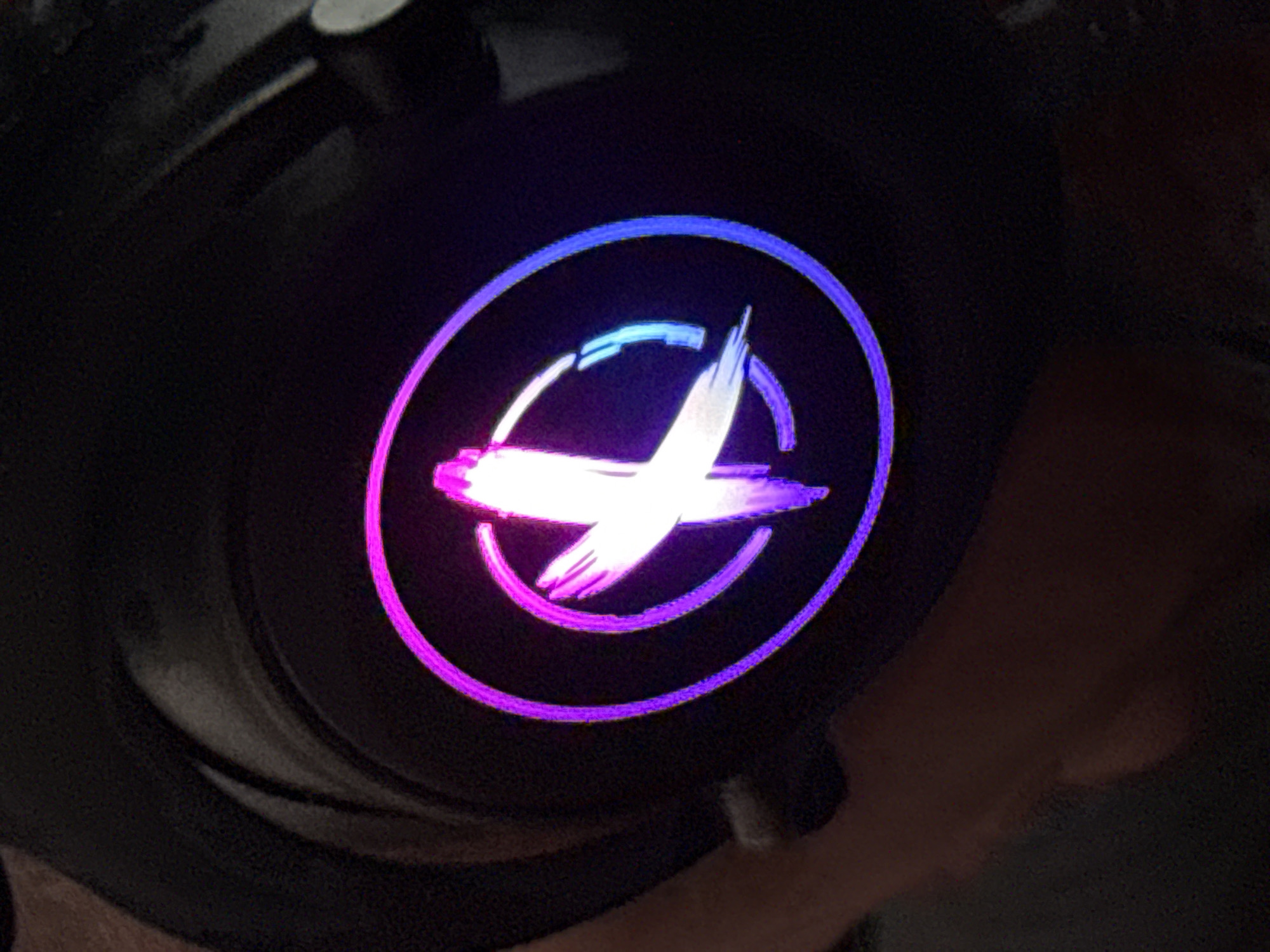 The media file is a close-up photograph of a black object with a circular, illuminated logo. The logo is in the center of the frame and features a stylized "X" shape formed by brushstrokes, all contained within a circular outline. The illumination of the logo transitions from pink and purple on the left side to a brighter white and blue on the right, with the outer circle also displaying a gradient of these colors. The object itself appears to be made of a dark, matte material, possibly plastic or metal. The background is dark and out of focus, with some indistinct shapes and textures that suggest an indoor, dimly lit environment. There are no people or animals visible in the image. The lighting on the logo is the primary source of light in the scene, creating a vibrant contrast against the dark background. There is no visible text in the image. The overall impression is that of a detail shot of a product, possibly a piece of electronic equipment or gaming hardware, given the common use of RGB lighting in such items. The focus is entirely on the illuminated logo, highlighting its design and color.