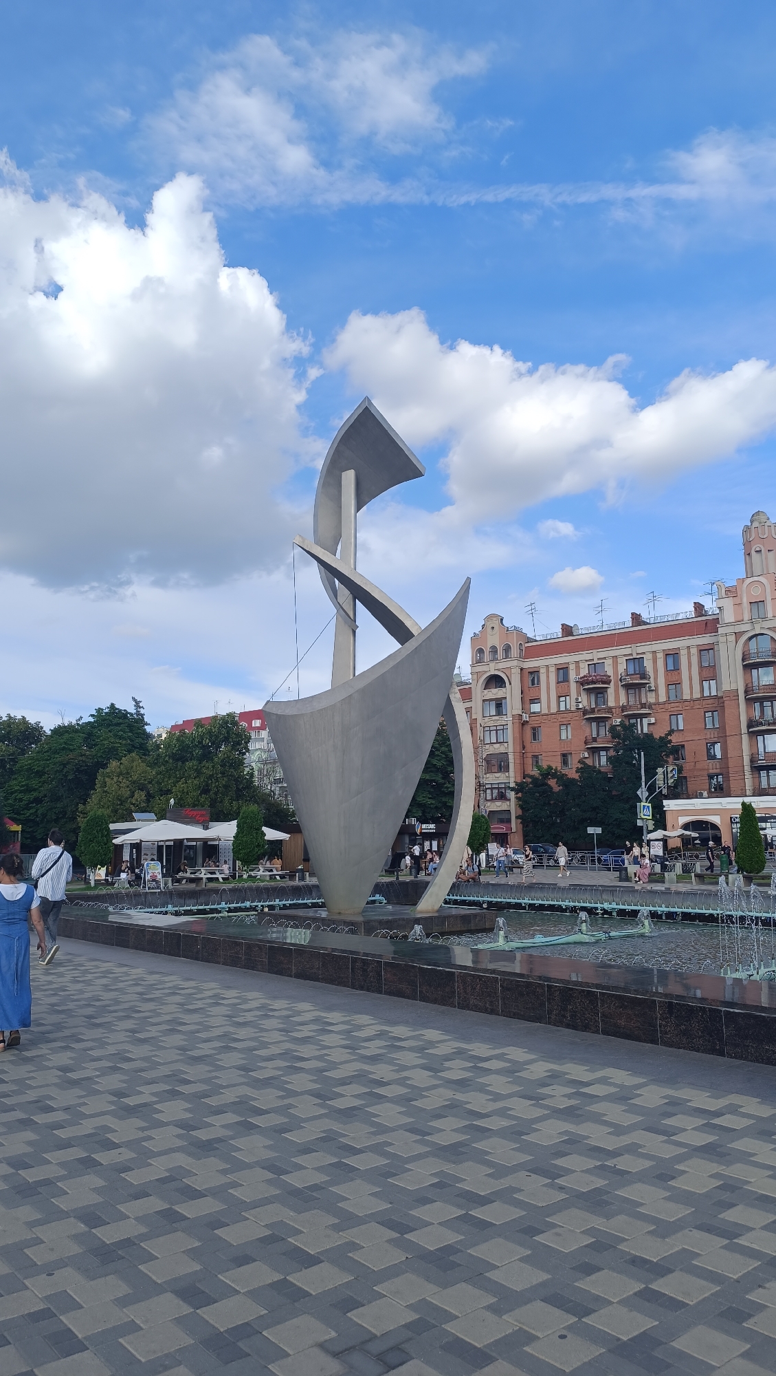 This is a wide shot of a public square in Novokuybyshevsk, Russia, on a partly cloudy day. In the foreground, a patterned gray-and-black tiled walkway leads towards a large, modern abstract sculpture made of gray metal. The sculpture resembles a stylized boat or a flame, with swirling, curved planes. Behind the sculpture is a large, rectangular fountain with multiple jets of water.
In the middle ground, there are several people walking along the walkway or sitting near the fountain. Beyond the fountain and sculpture are some trees and low buildings with white awnings that appear to be cafes or restaurants. Further in the background, there are multi-story apartment buildings with reddish-brown facades, typical of Soviet-era architecture. The sky is blue with scattered white cumulus clouds. The overall scene depicts a pleasant urban public space.