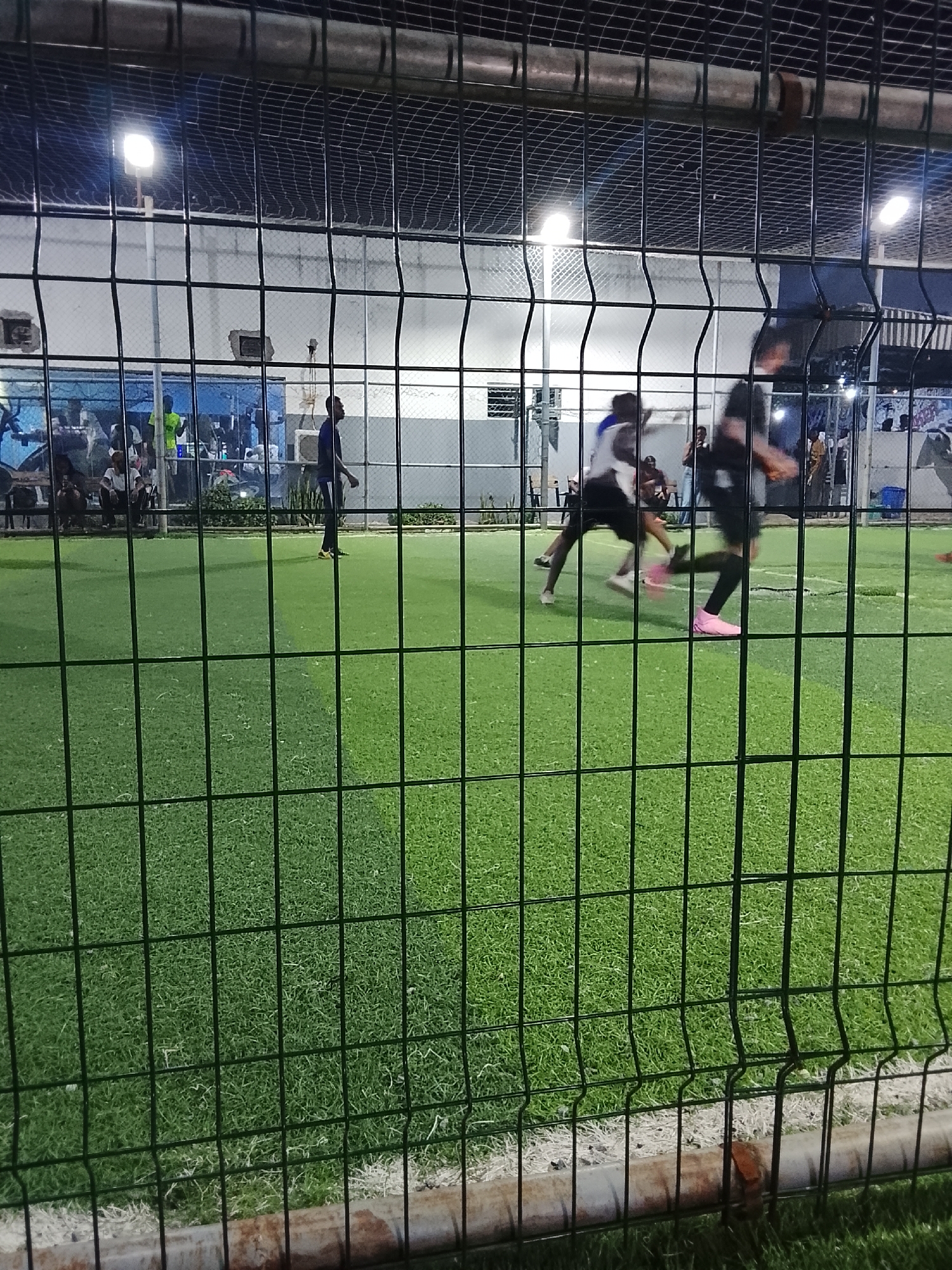 In Akwanga, Nigeria, a fenced outdoor artificial turf field is illuminated by overhead lights. Through the fence, several people play a game; two players appear to be in motion, one wearing pink footwear. Spectators are seated or standing beyond the field's perimeter. A low wall or barrier borders the field's edge where the fence is situated.