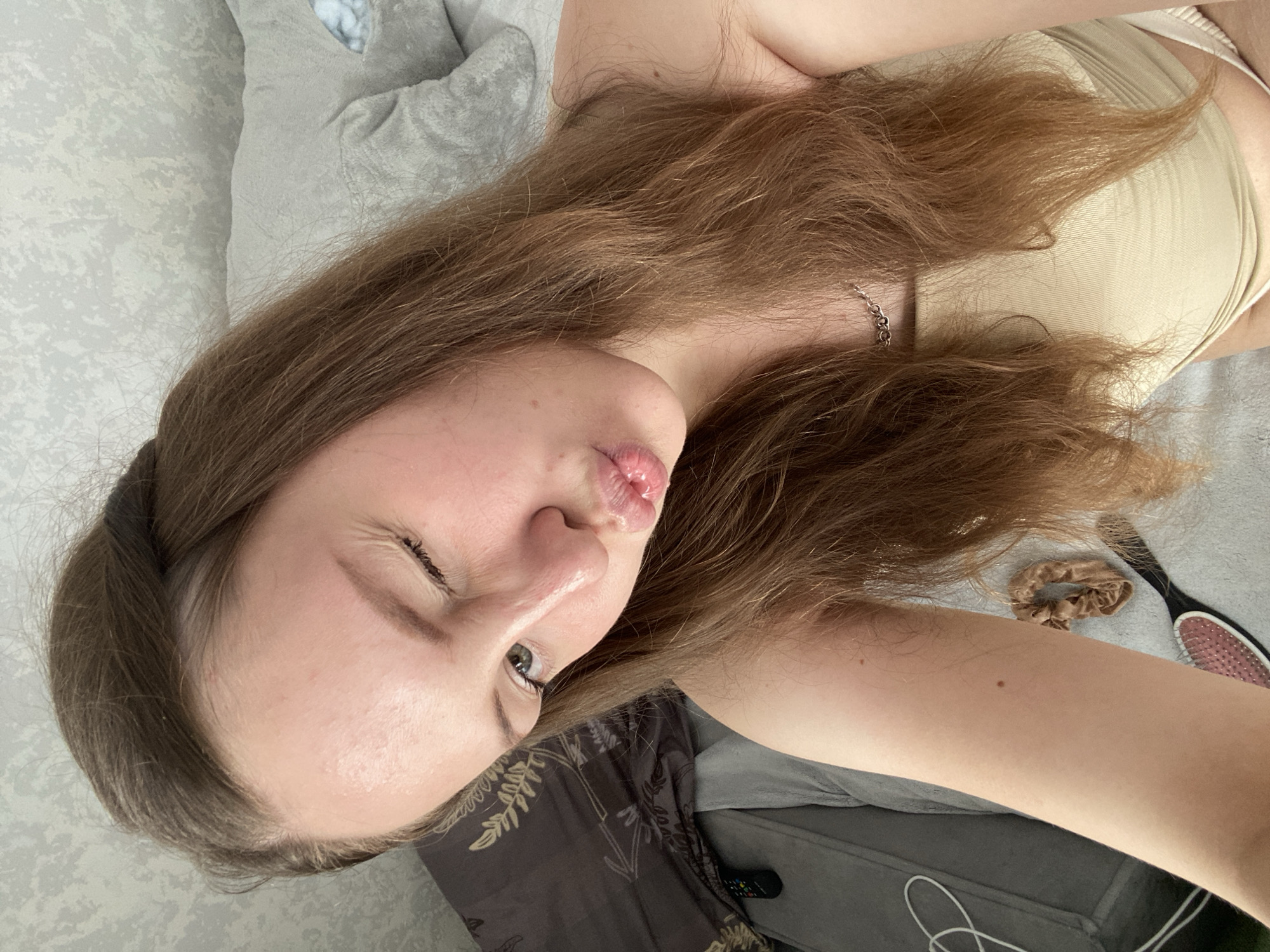 A young woman with long, light brown hair is lying down in bed, looking at the camera. She is winking and puckering her lips. She is wearing a light beige top. The background shows a patterned pillow and a gray headboard. A remote control is visible on the pillow. A hair tie and a hairbrush are on the bed in the foreground. The image appears to have been taken indoors, in a bedroom. There is no information in the image to suggest it is located in the city of Izhevsk, Russia.
