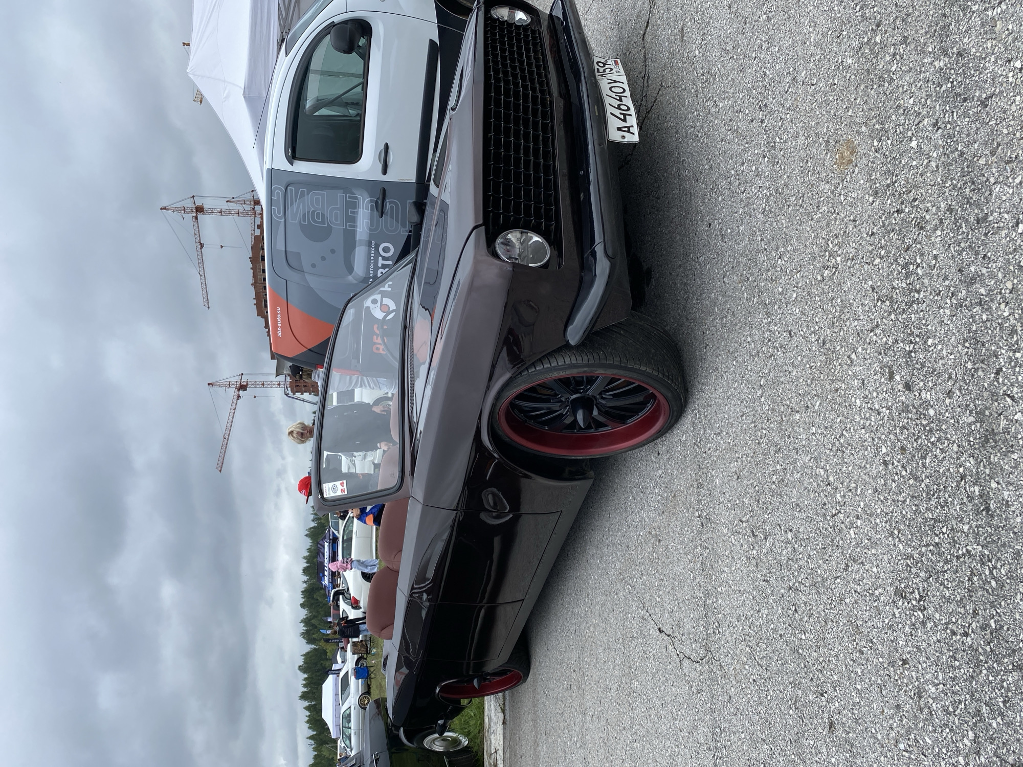 An outdoor, overcast medium shot captures a customized dark brown convertible parked on cracked asphalt. The car features prominent red rims with black spokes and a dark grille. A Russian license plate, reading "A4640Y 159", is affixed to the front. In the background, a white van with orange and gray graphics is visible. Several people are present, including a blonde individual with a red cap and another person wearing a pink hooded sweatshirt. Construction cranes and tents are also situated in the distance, suggesting an event or a construction site in the city of Perm, Russia.