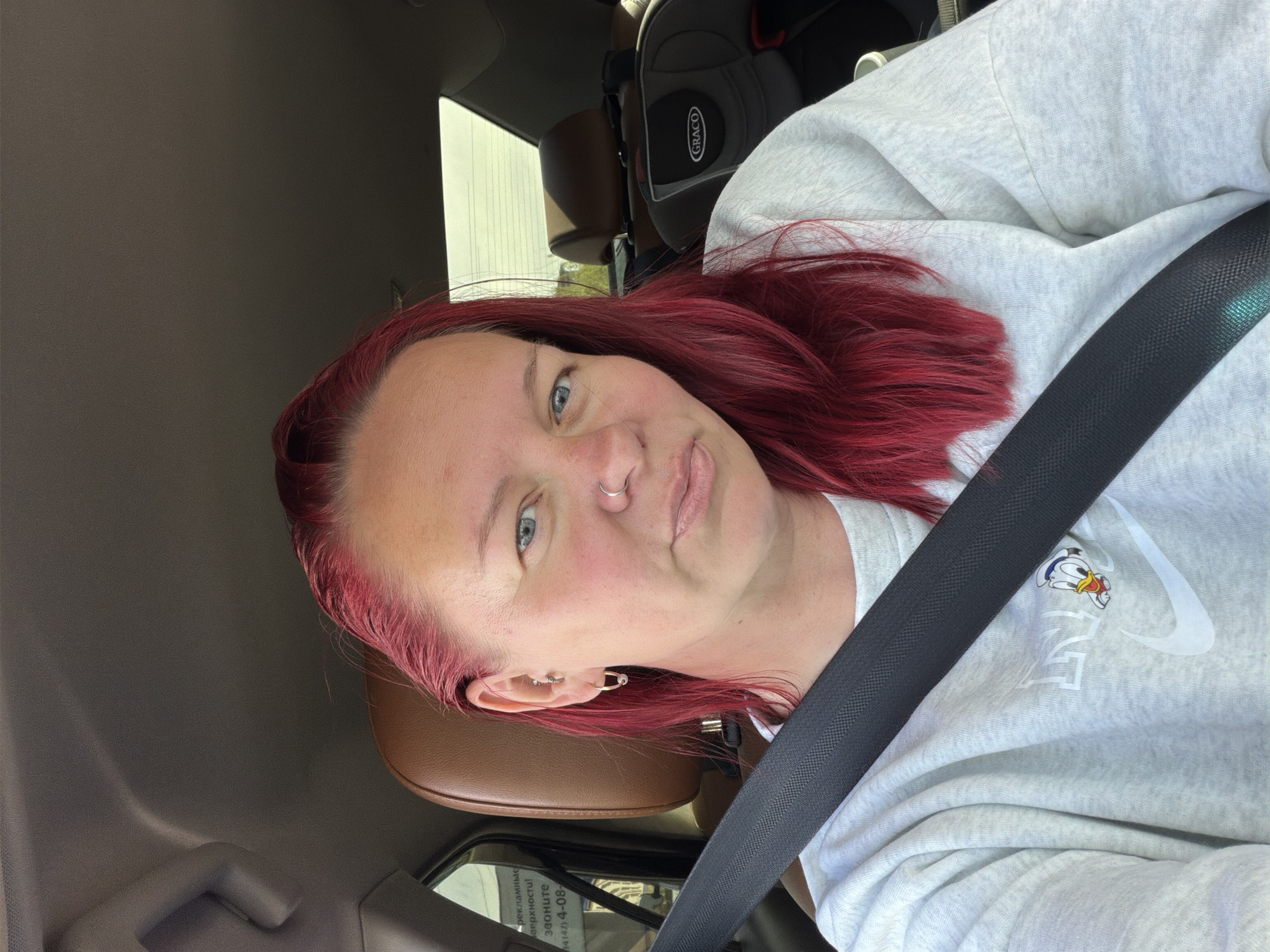 A person with red hair, wearing a light grey Nike sweatshirt with a Donald Duck emblem, is seated in the driver's seat of a vehicle. They are wearing a seatbelt. A Graco car seat is visible in the back. The vehicle's interior is brown. A glimpse of an exterior environment is visible through the car window, with Russian Cyrillic writing on a sign, and a phone number indicated (possibly 4-08-). Based on the Cyrillic sign, the location is Chaikovsky, Russia.