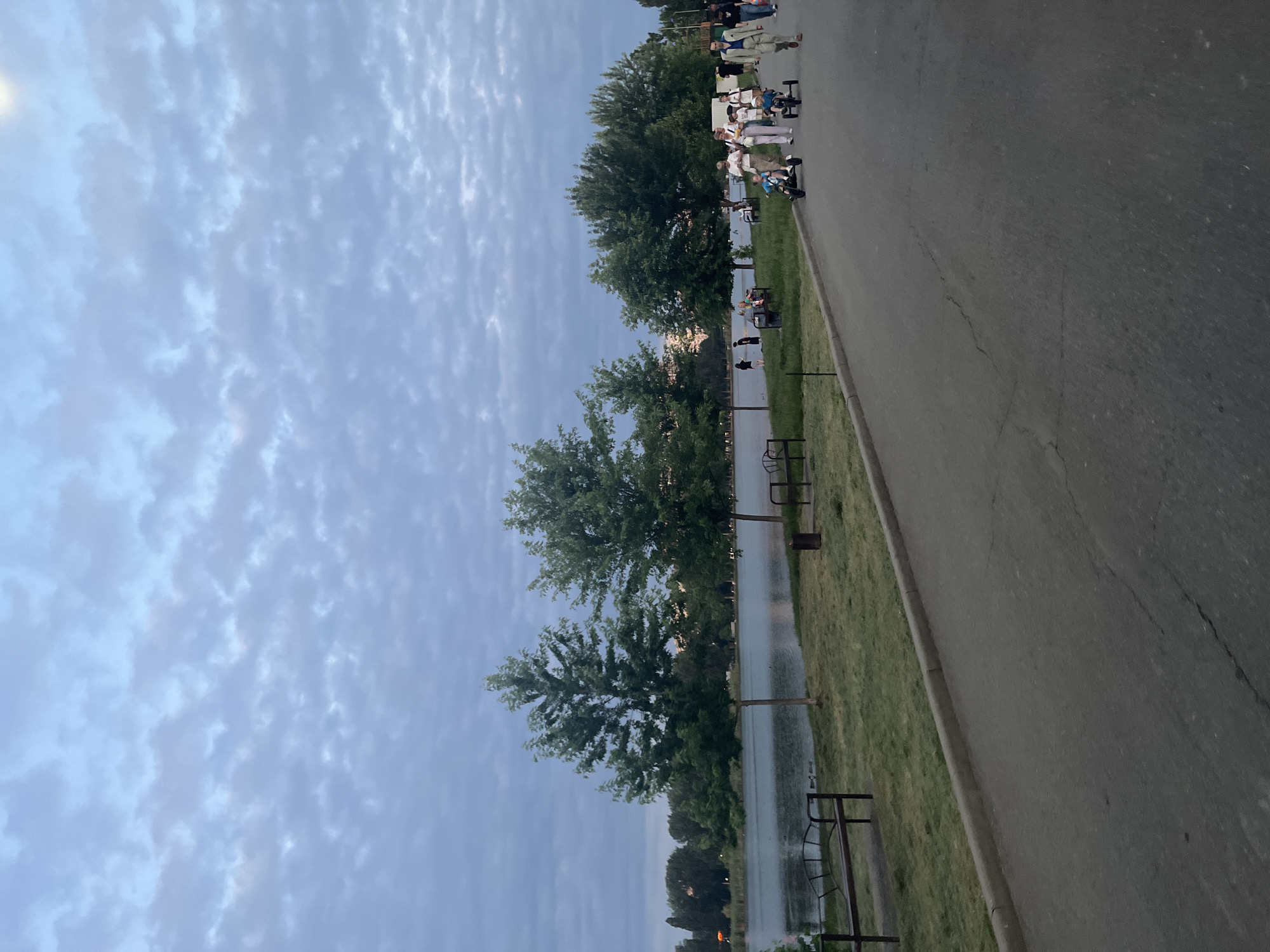 This is a full, eye-level, outdoor shot of a park next to a lake, likely taken in Krasnodar, Russia, during the late afternoon or early evening given the overcast sky and muted lighting. In the foreground, a paved asphalt pathway curves from the right side of the frame towards the left. Along the left side of the pathway is a strip of green grass, and beyond that, the calm surface of a lake. Several trees with full green canopies are spaced along the edge of the lake, with taller trees and more dense foliage visible further in the distance on the far side of the lake.
On the pathway, several people are visible. On the right side, a group of adults and children are walking. Two children are riding on small pedal-powered carts. One adult is pushing a child on one of these carts, while another adult walks beside them. Further down the path, more people are seen walking in the same direction. On the left side of the pathway, near the lake's edge, there are several benches. One bench is clearly visible in the left foreground, with another further down.
The sky is a soft blue-grey, filled with a dense, textured pattern of scattered clouds. The overall atmosphere of the image is peaceful and recreational, suggesting a popular spot for outdoor activities.