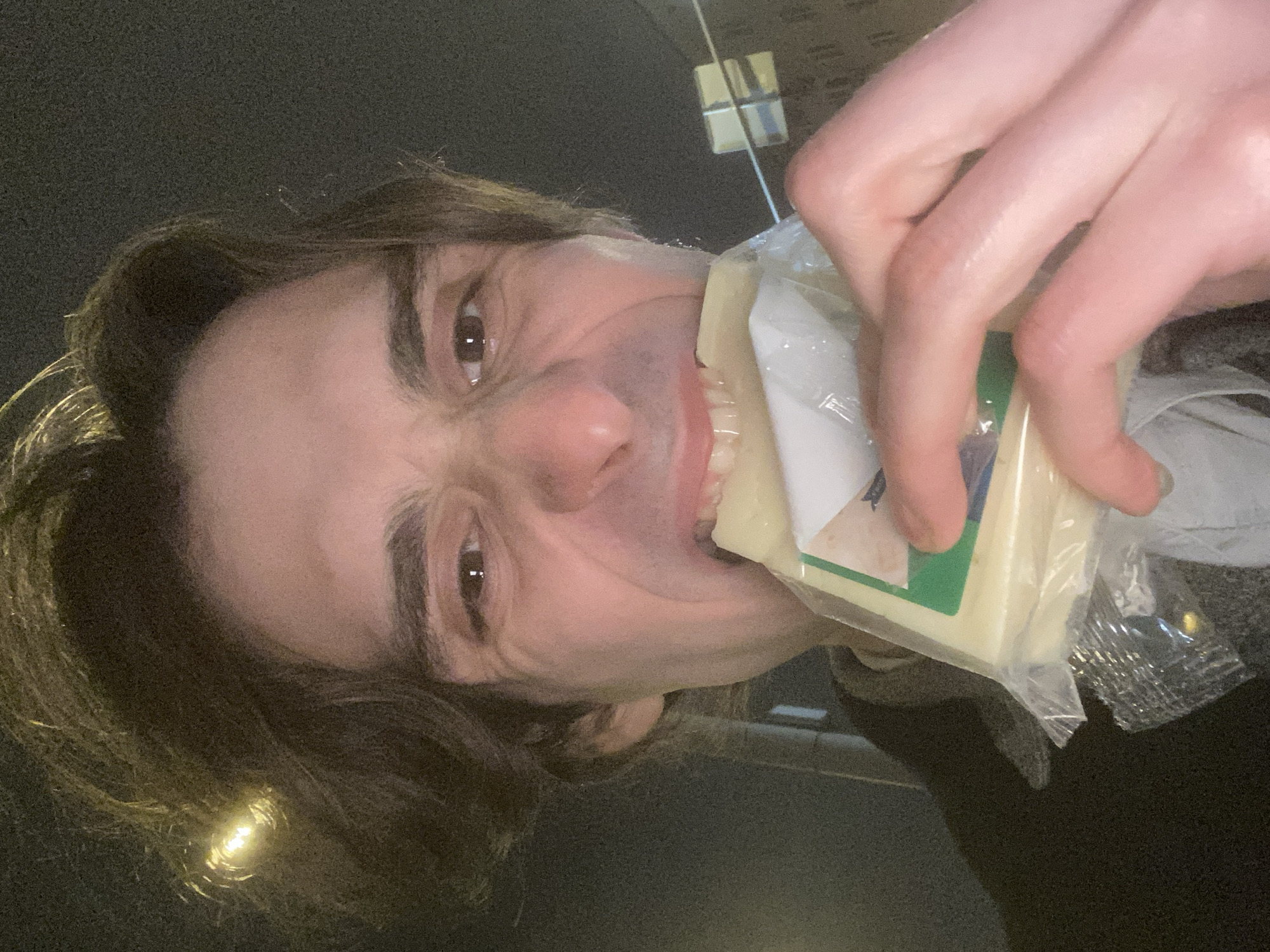 A male individual with brown hair and light skin is holding a packaged block of cheese in his mouth, with part of the cheese visible between his teeth. His face is angled upwards in a selfie pose, with a slight scowl. The background appears to be an urban setting, with a vertical rectangular structure topped by a horizontal feature, possibly architectural details or a light fixture. The packaging of the cheese is mostly clear, with a green and white label partially visible. The picture is taken with the camera tilted to the left. The location cannot be confirmed as Moscow, Russia based on the image content alone.