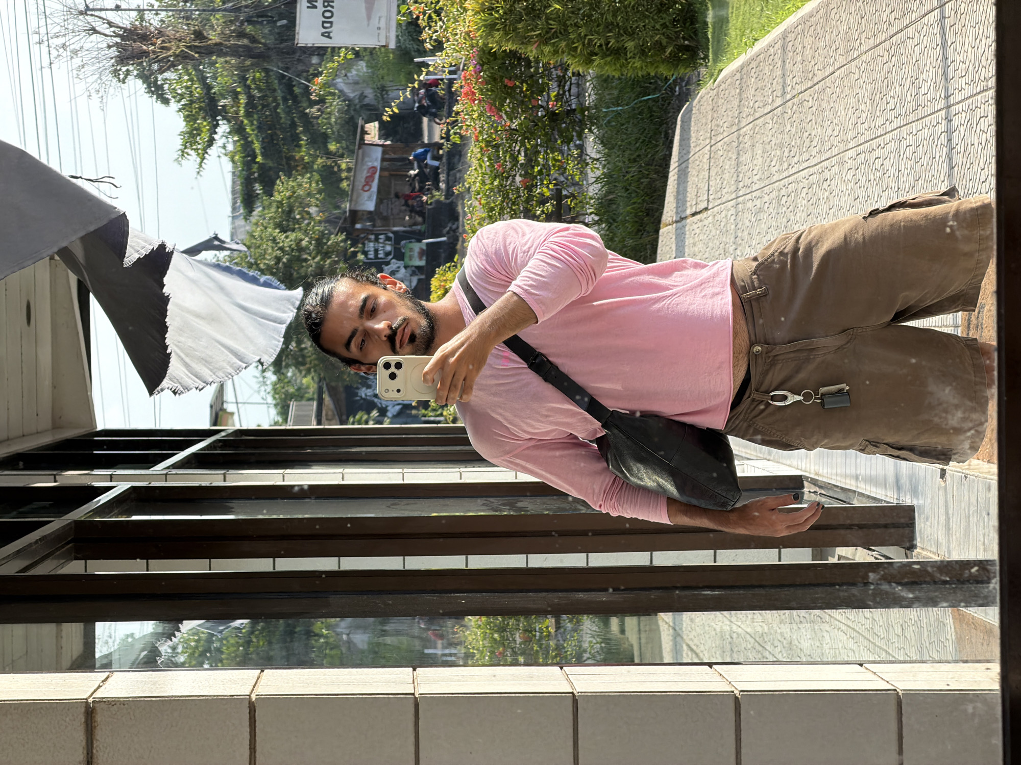 A man with dark hair and a beard is taking a mirror selfie in Tempurejo, Indonesia. He is wearing a pink long-sleeved shirt, brown cargo shorts, and a black crossbody bag. His phone is held up to take the picture, and a set of keys hangs from his shorts. He is standing on a paved walkway, with a building and lush greenery in the background. The scene appears to be outdoors during the daytime.