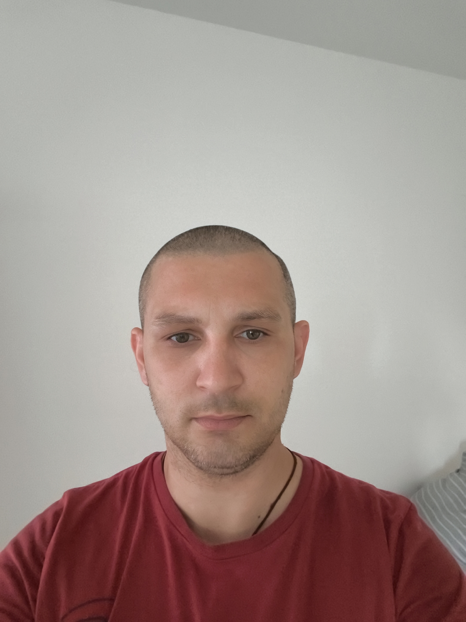 This is a portrait-style, medium-shot photograph of a light-skinned man with a shaved head and light stubble, wearing a dark red t-shirt. He is positioned centrally in the frame, facing the camera directly. The background is a plain, light-colored wall, likely an interior setting, as suggested by the presence of a striped fabric object to the right of the frame, resembling a comforter or pillow. No specific location in Wolgast, Germany can be determined from the image.