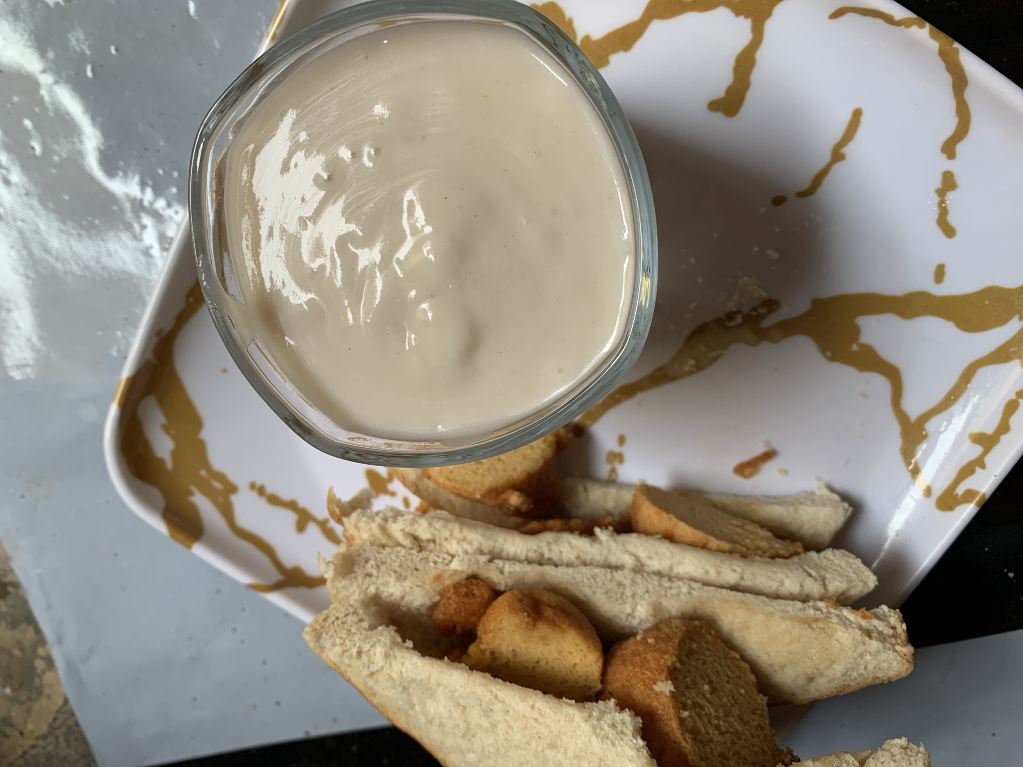 A top-down shot shows a white, rectangular plate decorated with gold paint strokes. On the plate sits a glass bowl filled with a beige, creamy sauce or dip. Next to the bowl are several open hotdog buns filled with chunks of fried, golden-brown bread. The setting is indoors, possibly a kitchen or dining area, and the image could be from anywhere, not necessarily Rafin Par, Nigeria. There are no people visible. The focus is on the food arrangement on the plate.