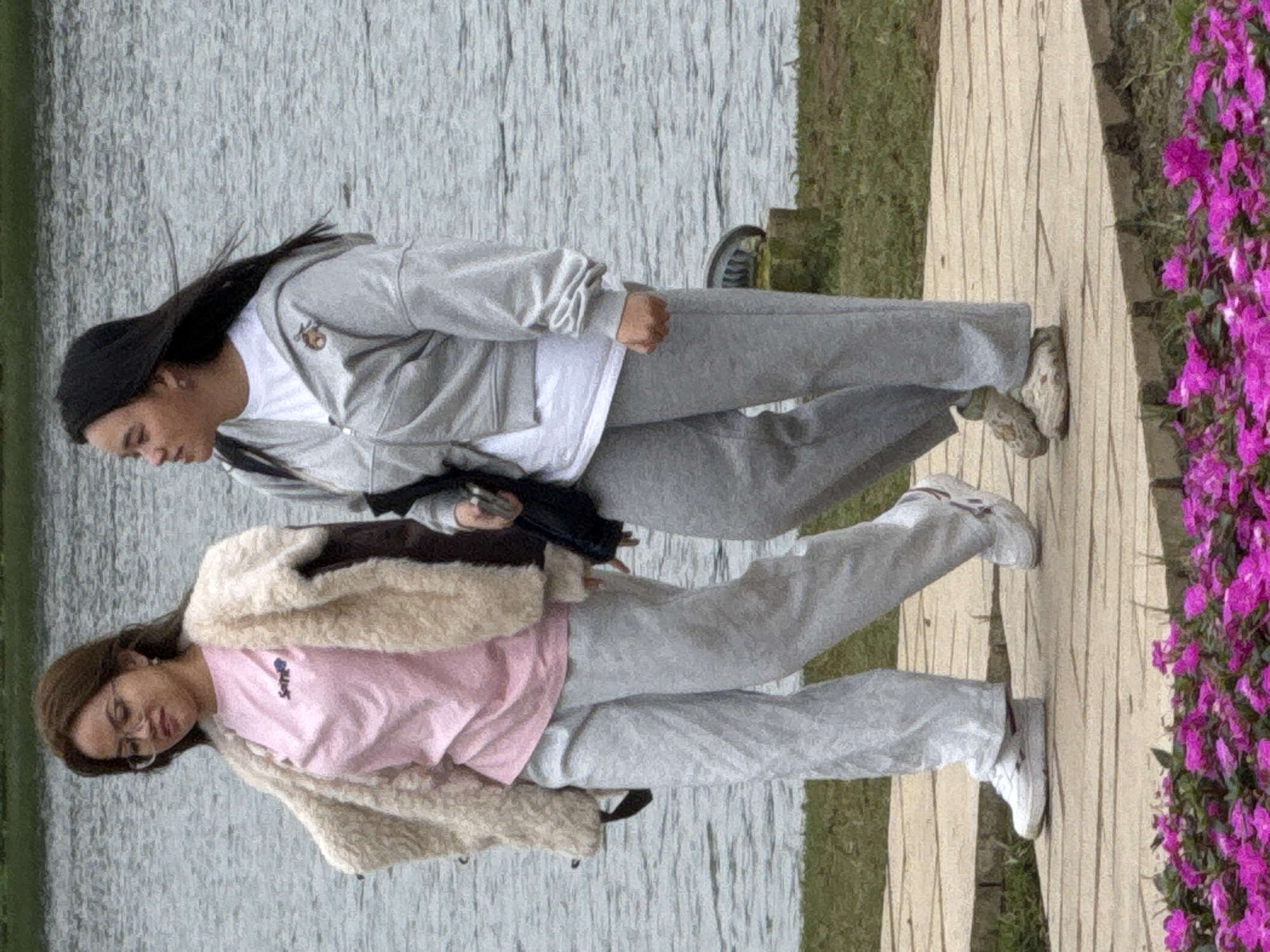 Two young women are walking on a paved pathway next to a body of water, likely a lake or river, in Da Lat, Vietnam. The woman on the left has brown hair and is wearing glasses, a pink t-shirt, a fluffy cream-colored jacket, and light grey sweatpants. The woman on the right has dark hair and is wearing a white t-shirt, a grey zip-up hoodie, and grey sweatpants. Both are wearing athletic shoes. The pathway is lined with green grass and a bed of vibrant pink flowers. The background shows a calm body of water.