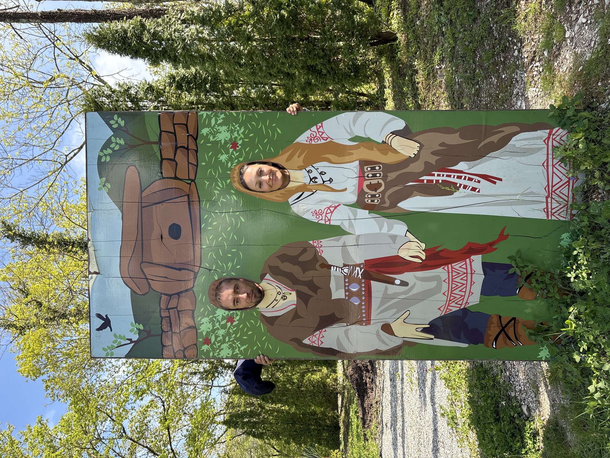 An outdoor scene features a large, brightly painted photo cutout board depicting two figures in traditional folk attire. Two individuals, a man and a woman, are posing behind the board, with their faces visible through circular cutouts. Both are smiling broadly, conveying a cheerful mood.
The board is crafted from wooden planks and showcases a female figure on the right with long, flowing brown hair, dressed in a white tunic with red embroidered patterns, a brown skirt, and a decorative brown belt. The male figure on the left has a beard and wears a white embroidered tunic, a brown vest or cloak, a striking red sash, and brown pants with criss-cross leg wraps. The painted background on the board includes green foliage, small red flowers, a large stylized brown wooden structure on the left, and a black bird in flight.
The actual background is a vibrant natural setting under bright daylight. Lush green trees, some with fresh spring leaves, stand tall against a clear blue sky with light clouds. The ground surrounding the board is a mix of gravel and green grass.
The activity involves the man and woman engaging in a fun photo opportunity, likely at a tourist attraction in Pshada, Russia. The time of day appears to be sunny, and the overall atmosphere is joyful. No visible text is present in the image.