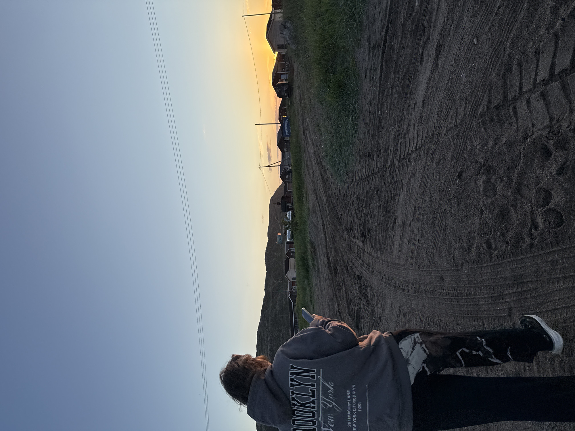 A person is standing on a dirt road in Teriberka, Russia, looking at their phone. The person is wearing a grey hoodie with "BROOKLYN New York" printed on the back, and black pants with a white marbled pattern. In the background, there are several small houses and hills. The sky is clear and blue, with a sunset in the distance. Power lines are visible stretching across the sky. The road has tire tracks from vehicles.