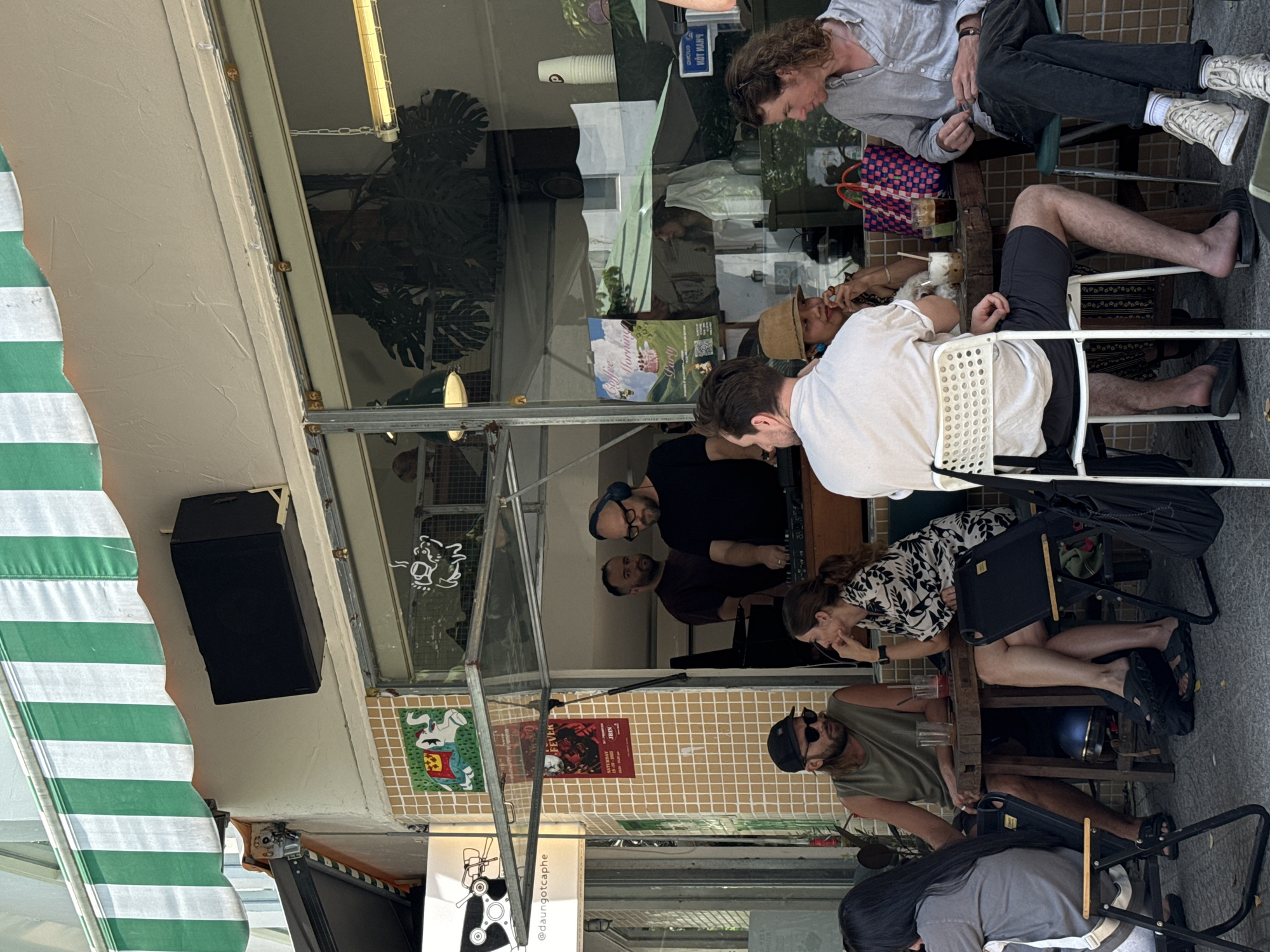The image depicts an outdoor café scene, likely in Đà Nẵng, Vietnam, judging by the signage "@daungotcaphe" (a Vietnamese café name) and general ambiance. Several people are seated at tables.
A man in a light grey shirt and dark pants sits at one table, while another man with bare feet sits across from him.
At another table, a man wearing a white t-shirt is seated, and a woman in a black and white dress sits near him. The woman looks toward another person. The man in a white t-shirt has a bag slung over his chair.
A man with a beard, wearing a black cap and sunglasses, is seated at a third table.
Visible in the reflection of the café window is a DJ booth set up inside. Two men are behind the DJ booth.
The café has a green and white striped awning. Posters and drawings are displayed on the wall below the window.