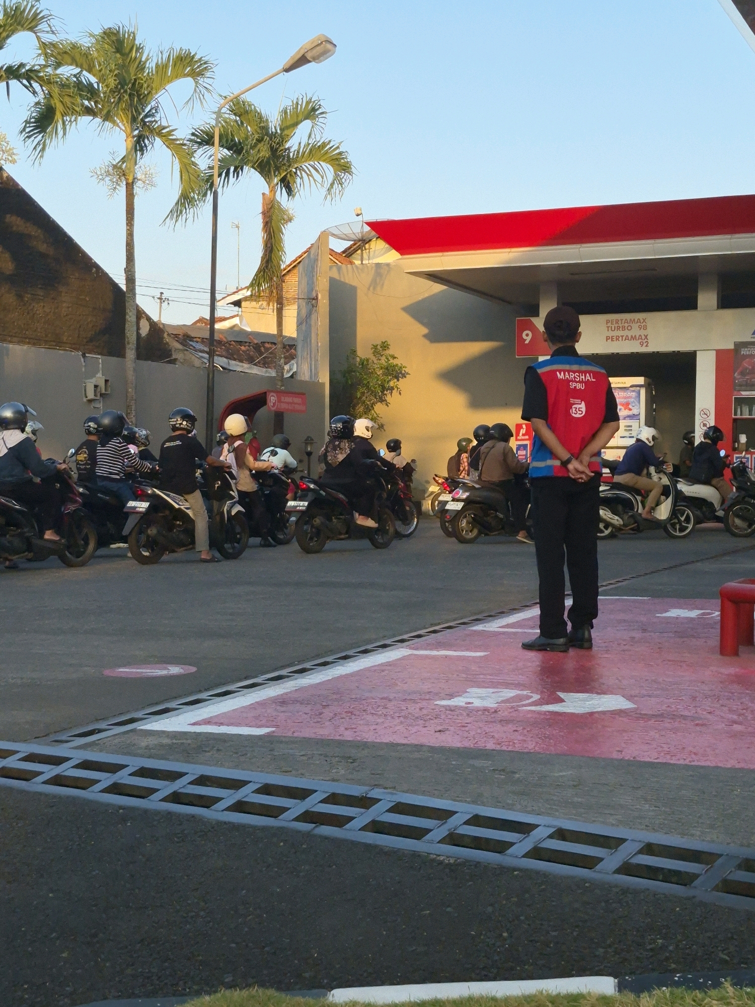 The image depicts a street scene at what appears to be a gas station in Purworejo, Indonesia. The sky is clear and blue, indicating it's likely daytime. Two palm trees are visible on the left side of the frame, with a street lamp positioned between them. A building with a red roof, typical of a gas station canopy, is prominent in the background.
In the foreground, a person wearing a red and blue vest with the word "MARSHAL" and "SPBU" printed on it stands facing away from the camera, looking towards the gas station. This individual is dressed in black pants and black shoes.
Several motorcycles are parked or moving in the scene. On the left, multiple individuals are seated on motorcycles, some wearing helmets. One person in the foreground on the left is wearing a white helmet and a dark jacket. In the middle ground, more motorcycles and riders are visible, with a concentration of them queuing or waiting.
The ground is asphalt with visible markings. A white and red painted area with arrows is present in the foreground, suggesting a designated lane or waiting area. A metal grate or drainage cover runs across the bottom of the image. The overall impression is a busy operational scene at a gas station, likely during peak hours.