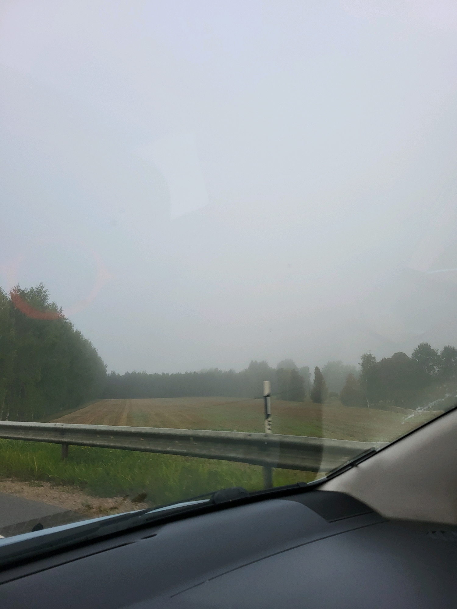 The image shows a view from inside a vehicle driving through a foggy landscape near Jūsmiņas, Latvia. The foreground is dominated by the dashboard of the car and the windshield, which has some streaks or water droplets on it. A metal guardrail runs along the side of the road, separating it from a grassy area and a field beyond. A line of trees can be seen on the left side of the frame, and in the distance, more trees and fields are obscured by dense fog. The sky is completely overcast and appears as a uniform light gray. There are no people or animals visible in the image. The overall impression is one of limited visibility due to heavy fog.