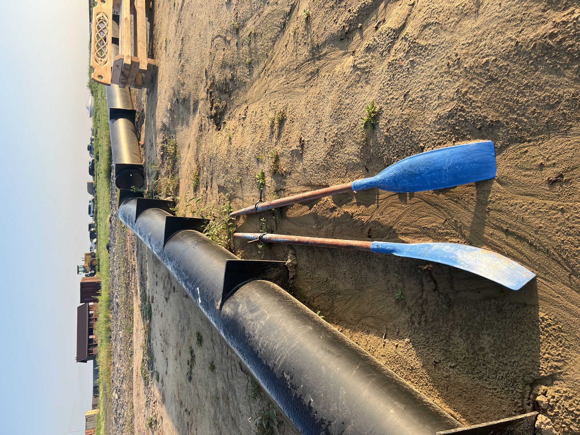 In Zav'yalovo, Russia, a construction site is depicted with a large black pipe running horizontally across the sandy ground. The pipe is supported by black metal brackets. Two blue-bladed paddles, one on the left and one on the right, are placed on the ground next to the pipe. In the background, several buildings, a yellow tractor, cars, and a white boat are visible under a clear blue sky. The environment appears dry and dusty with sparse green vegetation growing through the sand.