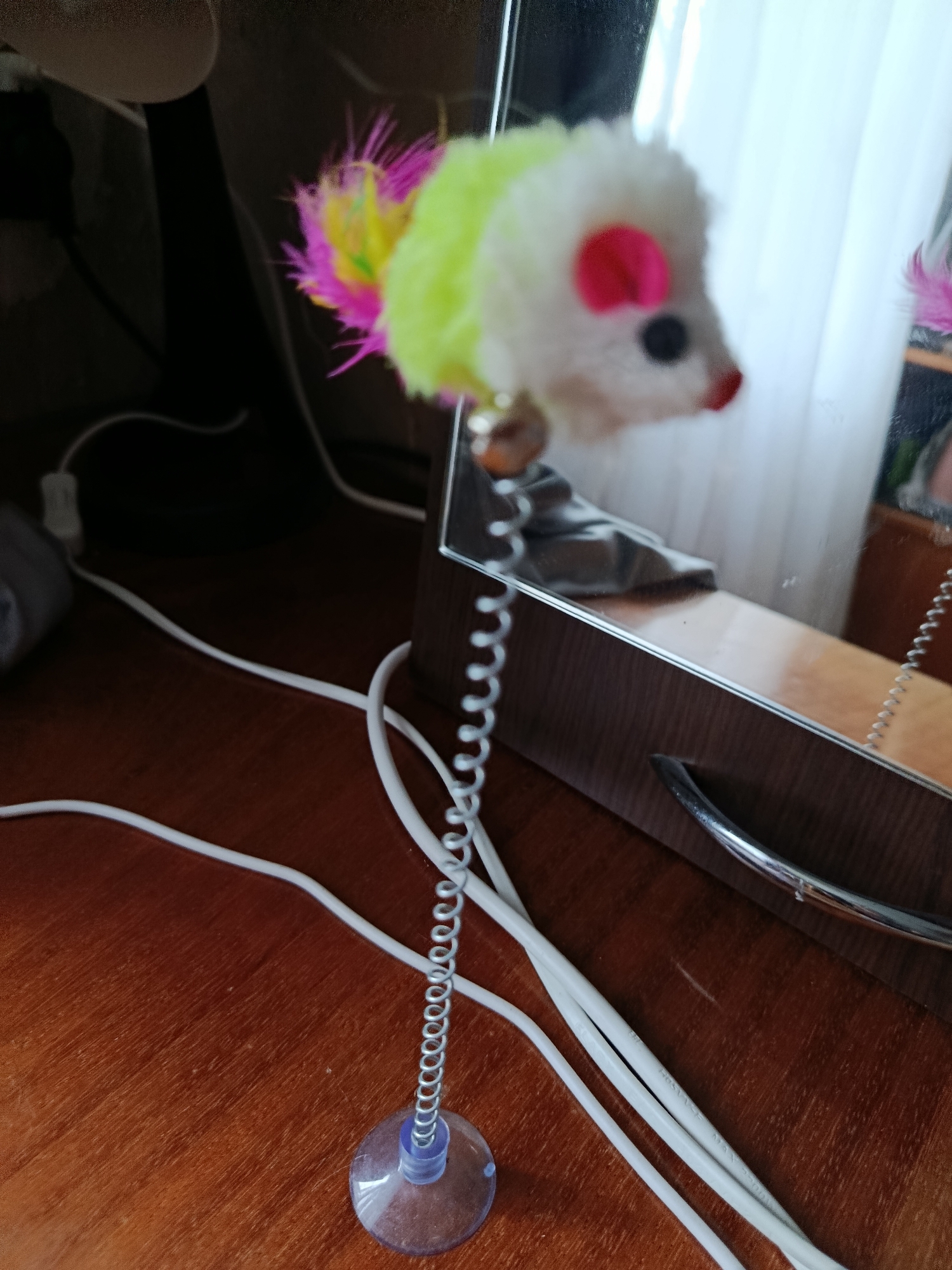 A toy mouse with a fluffy white body, yellow and pink feathers, and red ears is attached to a coiled spring, which is connected to a suction cup. The toy is positioned on a wooden surface near a mirror. Several white cords are visible on the wooden surface. The scene appears to be indoors, possibly in a domestic setting, with no observable connection to the city of Vladimir, Russia.