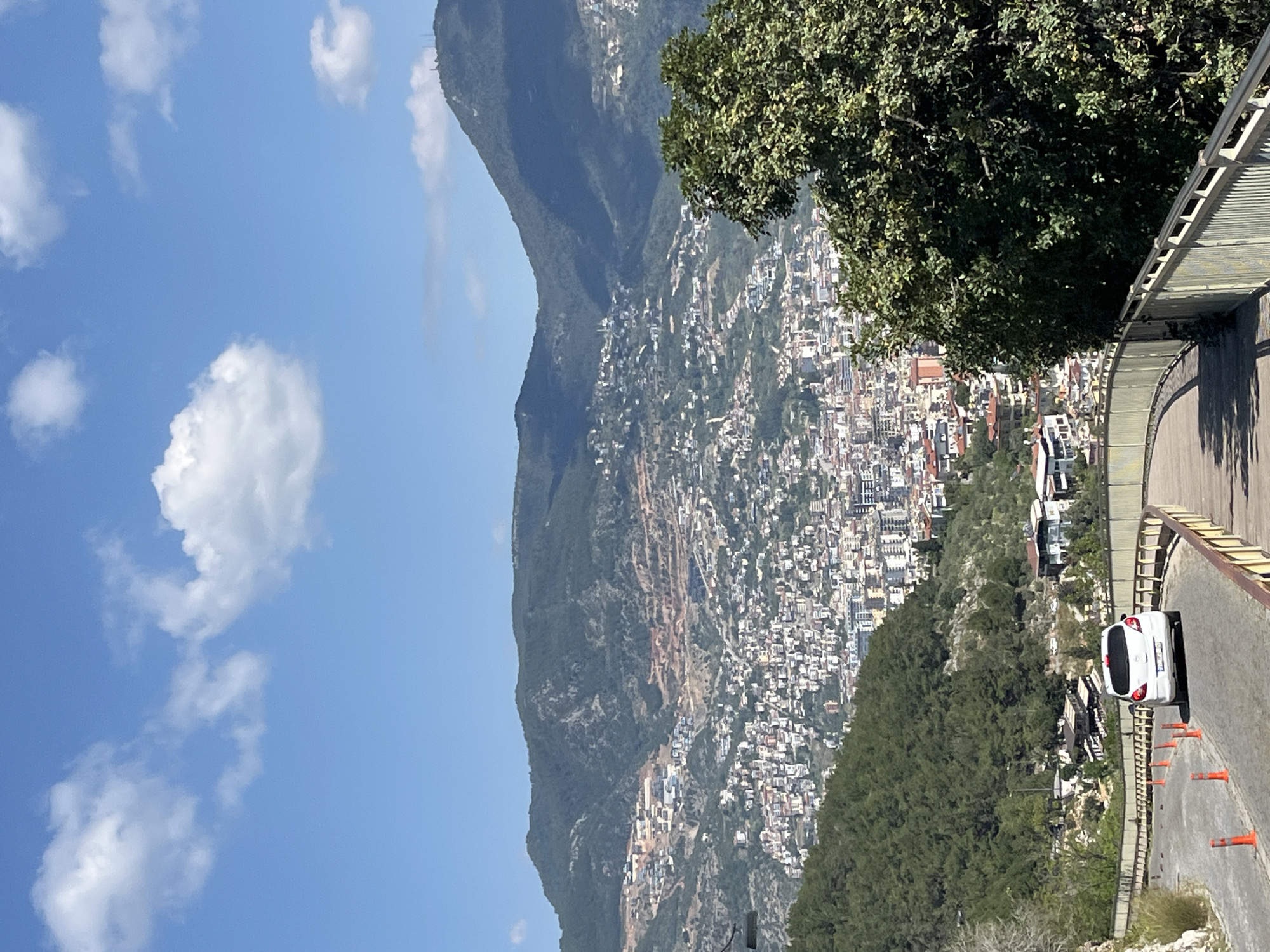 The image captures a high-angle view of a city nestled against mountains, likely Alanya, Turkey, given the geographical context. The sky, predominantly blue, features scattered white clouds. A mountain range forms the backdrop, with a dense urban sprawl cascading down its slopes towards the foreground. The cityscape consists of numerous buildings, predominantly white or light-colored, closely packed together. A road curves into the frame, with a white car parked alongside. Orange traffic cones are positioned on the road, suggesting some form of roadwork or temporary barrier. A tree with dense green foliage occupies the top right of the frame, partially obscuring the view. A metallic barrier runs alongside the road, indicating a viewpoint or overlook. The general atmosphere suggests a bright, sunny day.