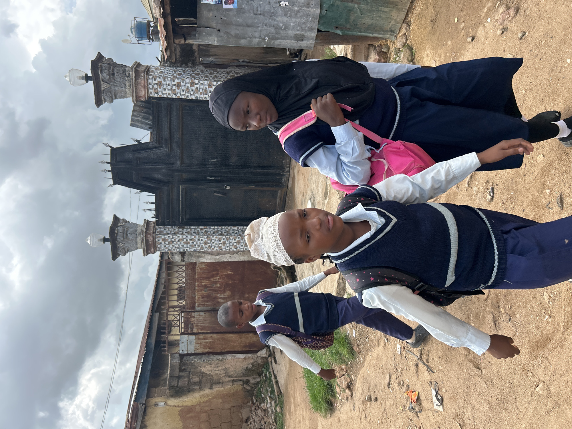 This outdoor image, taken in Jos, Nigeria, features three children in school uniforms standing on a dusty path under a cloudy sky. In the foreground, a boy wears a white long-sleeved shirt, a navy blue V-neck vest with white stripes, navy blue pants, and a white lace cap. He carries a backpack with a purple patterned strap and looks calmly towards the viewer.
Behind him and slightly to the left, a girl is visible, wearing a black hijab, a light blue shirt, a navy blue V-neck vest, and a navy blue skirt or dress. She carries a bright pink backpack and looks off to the left. Further back, on the left side of the frame, a younger boy in a similar uniform and a backpack looks straight ahead, not at the camera.
The background is characterized by an imposing dark metal gate, possibly part of a mosque or significant building, flanked by two ornate concrete pillars, each topped with a lamp fixture. To the left of the gate, weathered and rusty corrugated metal sheets form part of a fence or building. Beyond these structures, various buildings with corrugated iron roofs are visible, along with a blue water tank on a distant rooftop. The ground is dry, light brown soil with scattered debris, including small patches of green grass. The overcast sky indicates daytime. No discernible text is clearly visible on any signs or posters. The scene captures a moment of daily life, likely involving the children going to or from school, within an urban setting.