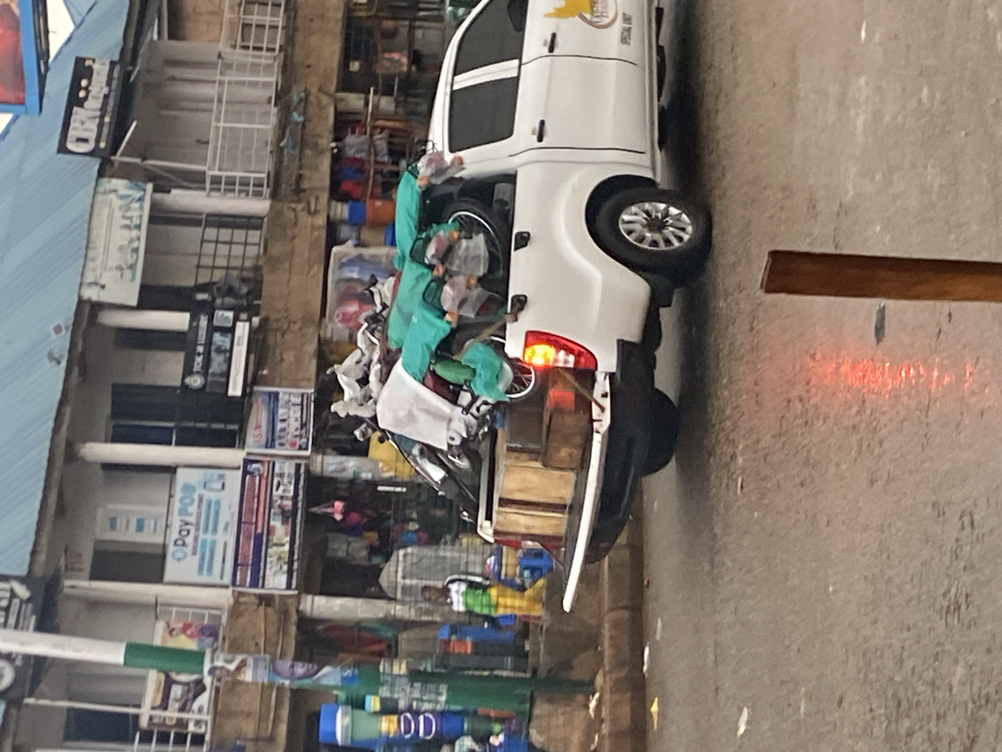A white pickup truck is loaded with several motorcycles and other items in what appears to be the city of Bukuru, Nigeria. The truck is parked on a street with a sidewalk lined with shops and market stalls. Some of the shops have signs with text in English and what might be local languages. One sign reads "PAY POS" with a logo that appears to be related to payment processing. The truck's bed is covered with a green tarp, and several motorcycles are visible underneath it, some strapped down. There are also wooden crates and other assorted goods piled in the truck bed. A person wearing a green and yellow outfit is standing on the sidewalk near the truck. The street appears to be wet, possibly from recent rain, and there is some debris scattered on the ground. The overall scene depicts a commercial area with significant activity, likely related to transportation or delivery of goods.