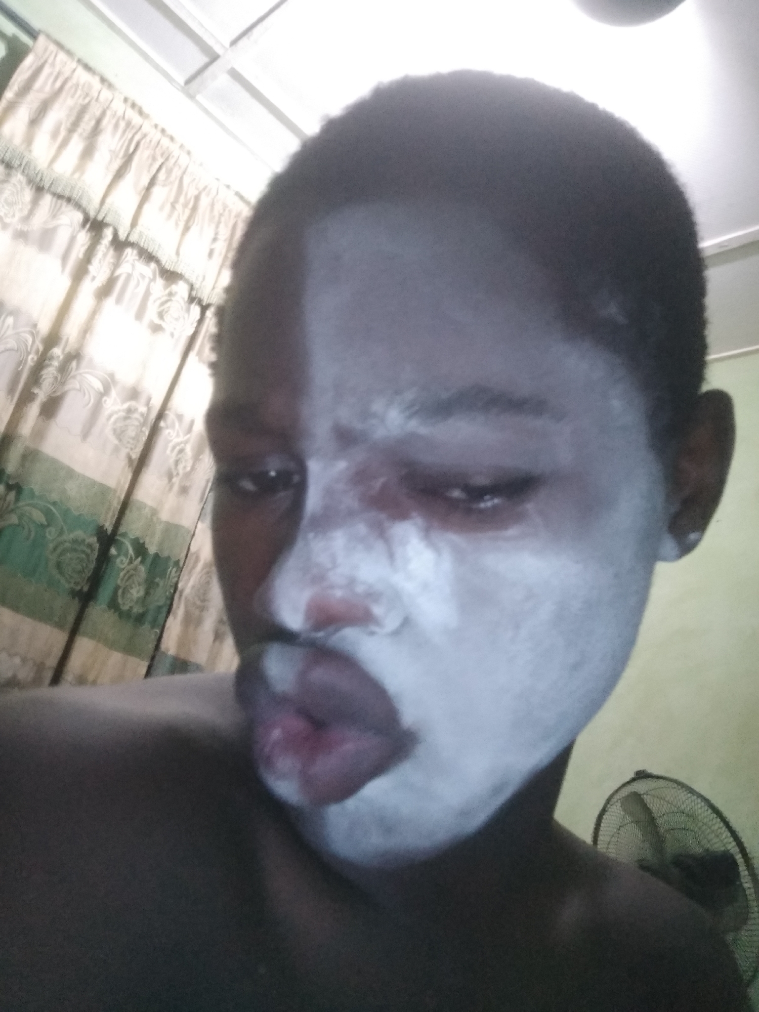 A close-up shot features a person with dark skin, approximately half of their face covered in a thick white substance. The person's mouth is puckered, and one eye is slightly squinted. The background includes a curtain with a floral pattern and a household fan. The location is unknown.