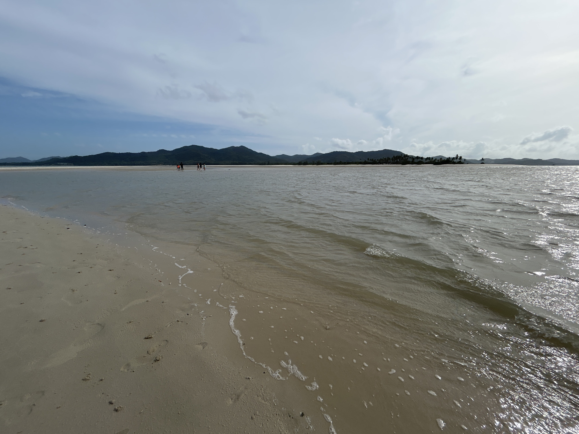 The image depicts a wide, shallow body of water extending towards a distant, low-lying mountainous coastline under a cloudy sky. A sandy beach occupies the foreground on the left, with footprints visible in the wet sand at the water's edge. Several people are walking in the shallow water in the middle ground, some appearing to be clustered together. The water is murky brown and shows ripples, with sunlight glinting off the surface. The coastline is covered in dark green vegetation and palm trees, with the mountains in the background appearing in various shades of blue and green. This scene is consistent with a coastal area on Koh Yao Noi or Koh Yao Yai, islands in the Phang Nga Bay, Krabi Province, Thailand, known for their shallow waters and tidal flats.