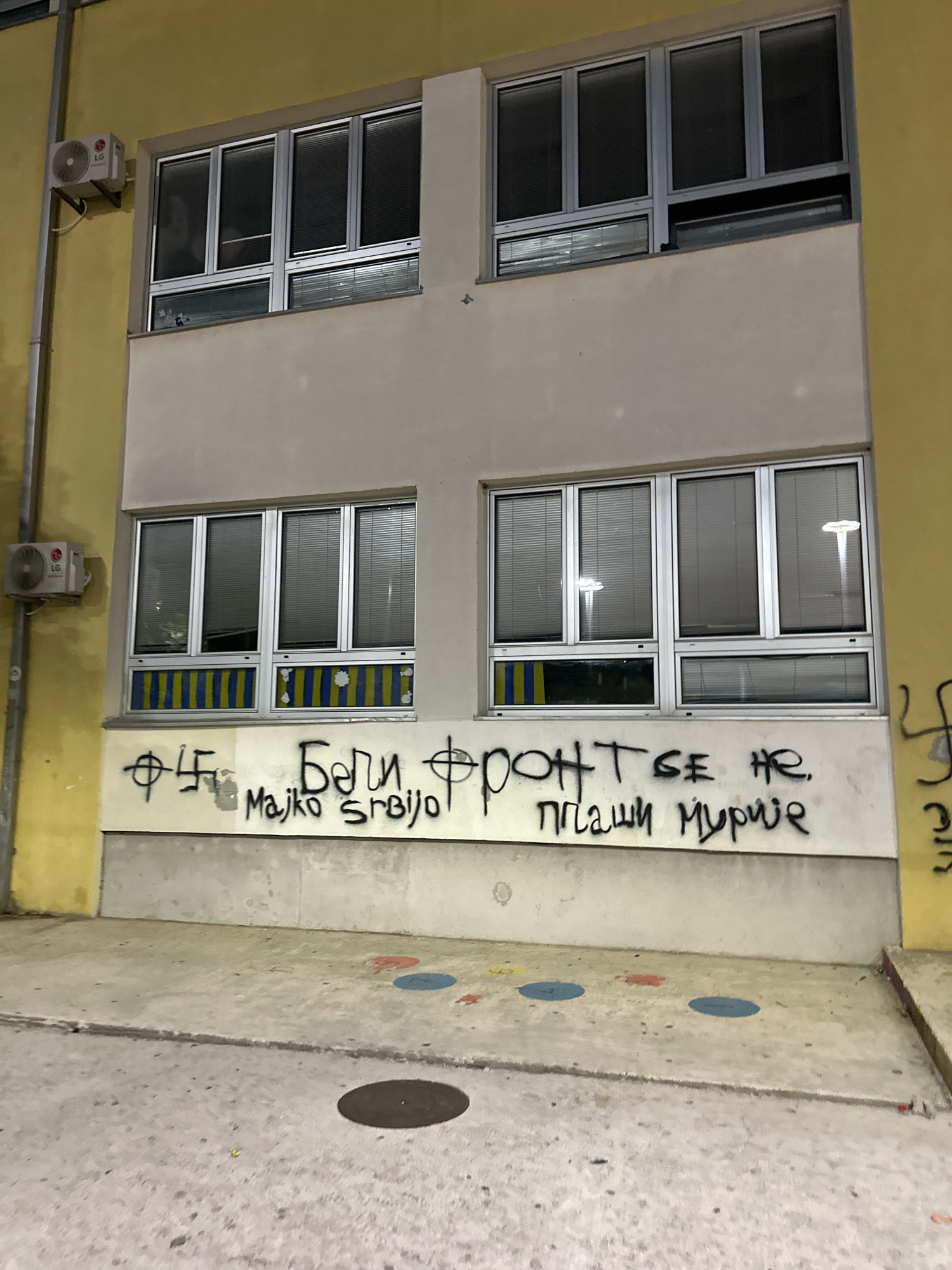 The image shows the facade of a building in Budva, Montenegro, with graffiti on its lower section. The graffiti includes a swastika symbol, Cyrillic script, and what appears to be the phrase "Beli front se ne plaši Murinje" which translates to "The White Front is not afraid of Murinje". Below this, the text "Majko Srbijo" is visible, meaning "Mother Serbia". The building has several windows with blinds and is painted in yellow and beige colors. There are also two air conditioning units mounted on the exterior wall. The ground in front of the building is a concrete walkway with painted colorful circles. The overall scene suggests a public outdoor space, possibly urban, with evidence of vandalism.