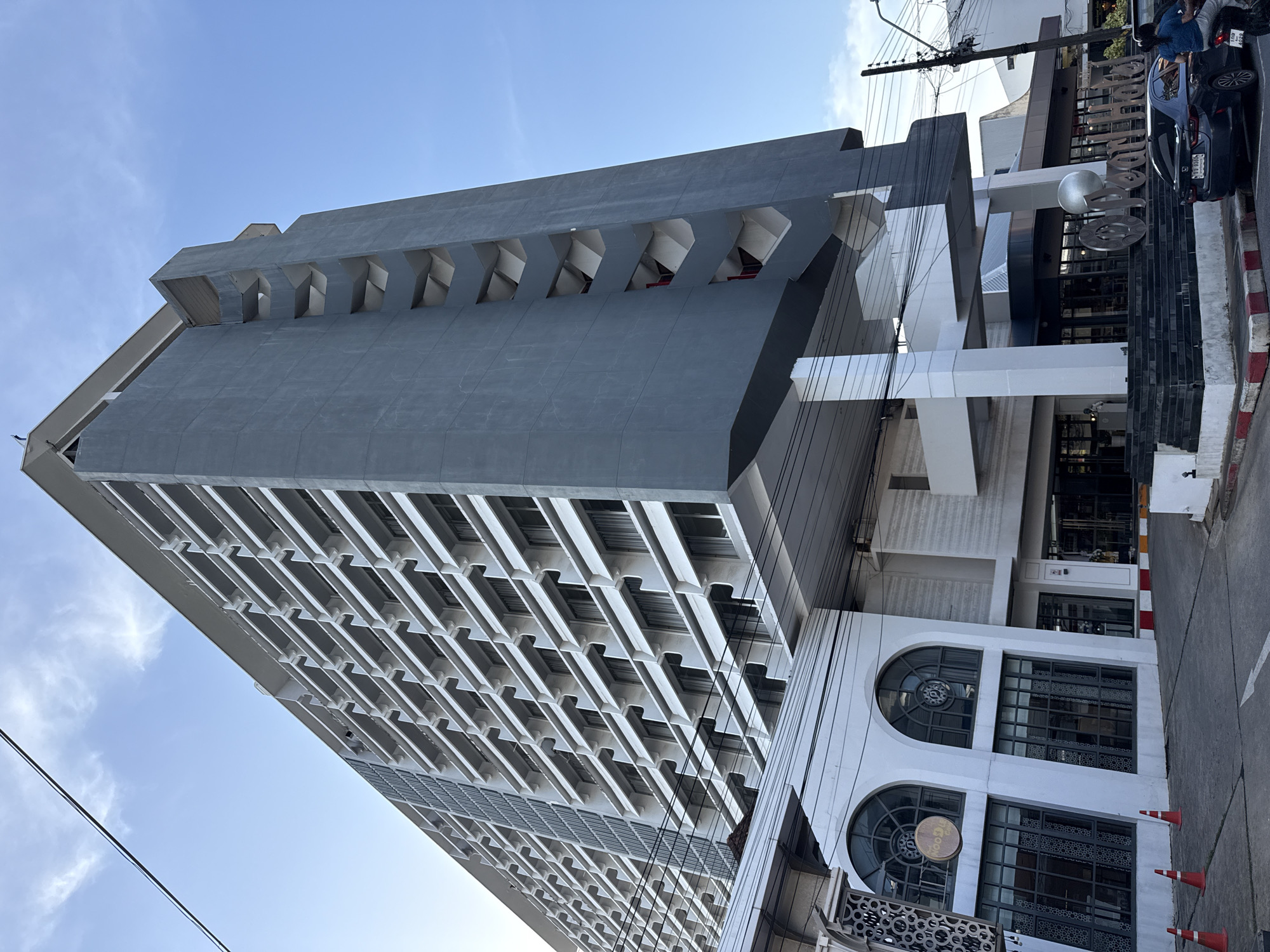 In Phuket City Municipality, Thailand, an upward-angle shot captures a modern, imposing hotel building with a distinctive gray facade. The hotel features a grid of white balconies and windows that ascend many stories, contrasting with the gray concrete. A unique architectural element is a series of triangular cutouts on the gray side of the building, revealing glimpses of what appear to be stairwells with red accents.
In the foreground, the lower section of the building displays a white, curved facade with large arched windows and a sign that reads "Noodle Cafe." The street level shows a paved road with traffic cones and a white directional arrow painted on the asphalt. A dark-colored car and a motorcycle with two occupants are parked near the entrance of the hotel, which is marked by a prominent "Ocean Hotel" sign with large metallic letters. Electrical wires are visible overhead, crisscrossing the frame against a clear blue sky with wispy clouds. The scene is likely captured during daylight hours.
