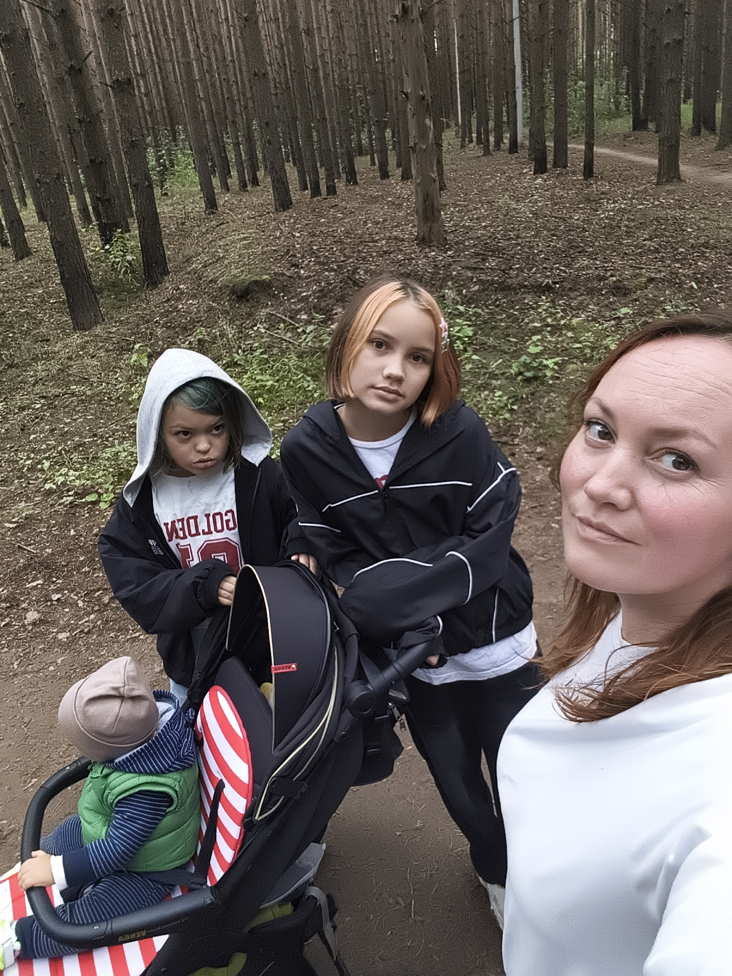 A group of four individuals, three children and one adult woman, are posing for a selfie in a wooded area. The woman, in the foreground on the right, is looking towards the camera. To her left, a teenage girl with reddish-blonde hair and a black jacket is also looking forward. Next to her, a child in a gray hooded jacket with green-tinted hair is looking down. In front of these two children is a stroller containing a baby wearing a green vest and a striped outfit. The background consists of a dense forest with tall, thin trees and fallen leaves on the ground. The path appears to be made of dirt. The overall scene suggests an outdoor recreational activity. The image was taken in Chaikovsky, Russia.