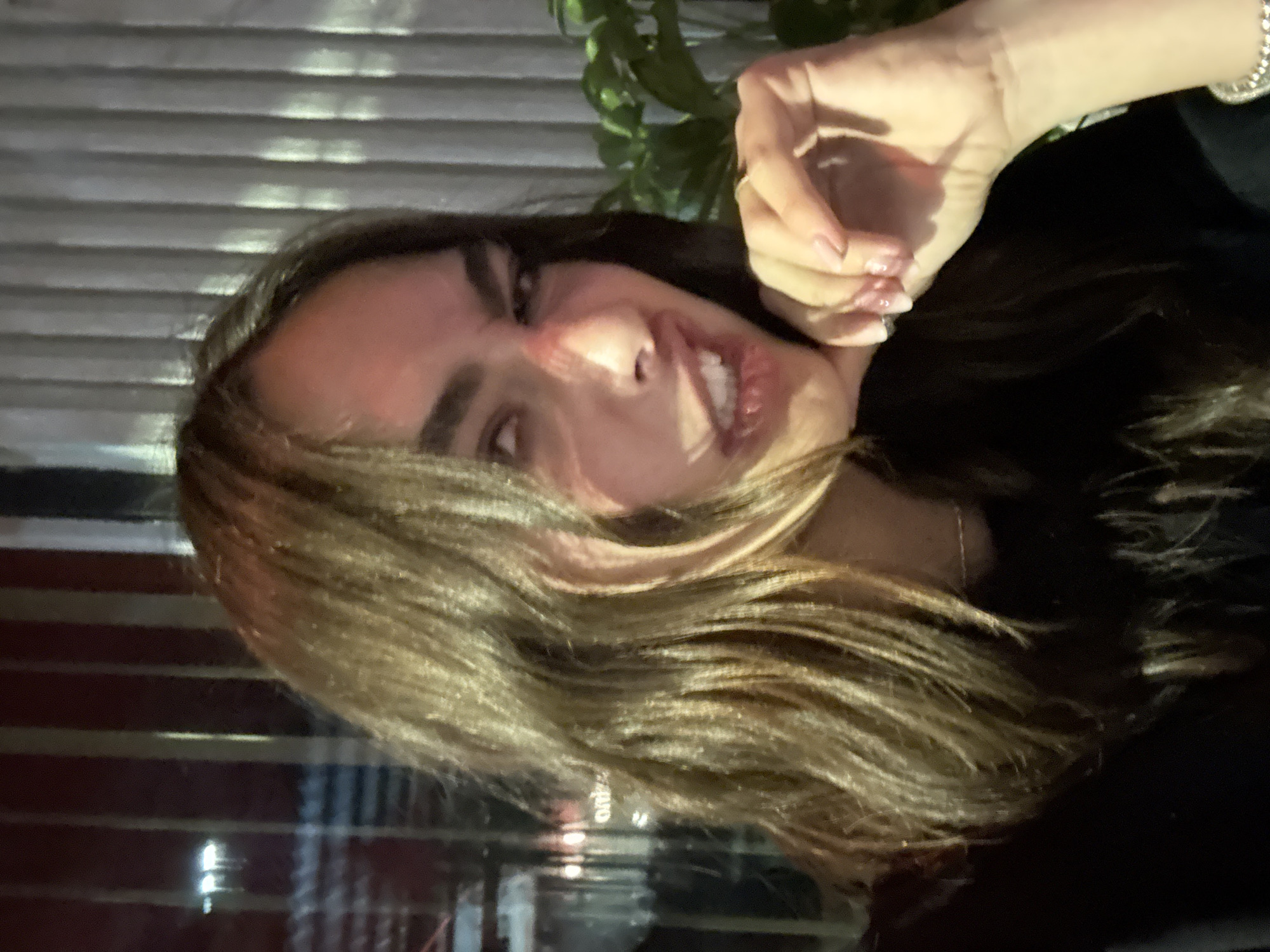 A young woman poses with her hand under her chin. She is standing indoors, in front of a window. Through the window, part of a red wall and white horizontal blinds can be seen. Behind the woman's head, some leaves are visible.