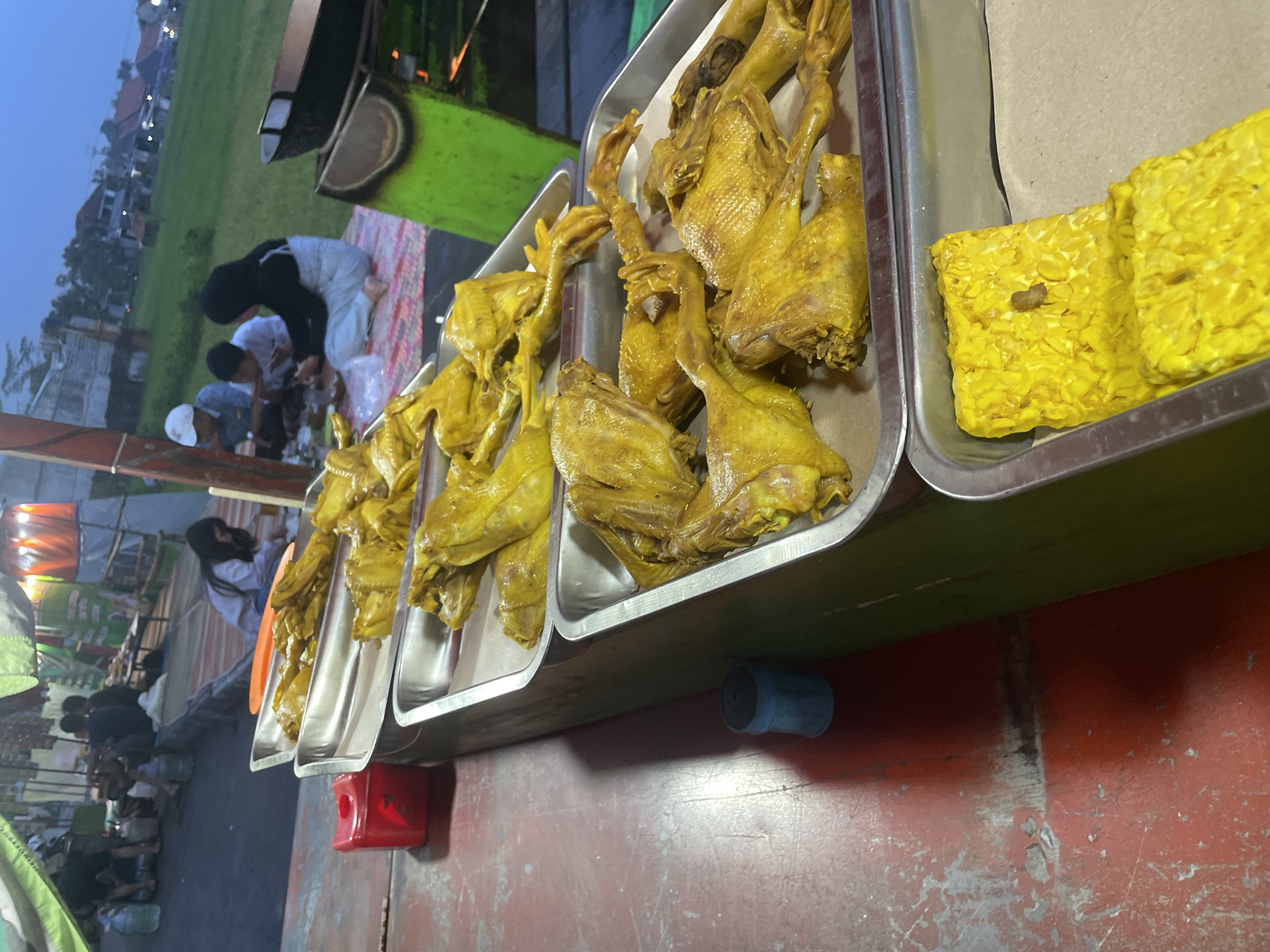 This image depicts an outdoor food stall, likely in Tasikmalaya, Indonesia, as suggested by the context of the food preparation. In the foreground, two metal trays are visible. The upper tray contains several whole, cooked chickens, uniformly yellow in color, presented as if ready for sale. The lower tray holds several rectangular blocks of fried tempeh, also yellow. Behind the food stall, a group of people are seated on a mat. Three individuals, two adults and a child, are actively engaged in eating. Further in the background, a few more people are tending to the stall or observing the scene. The environment suggests a casual outdoor setting with a mix of food preparation and dining areas. The overall lighting indicates it is either dusk or early evening.