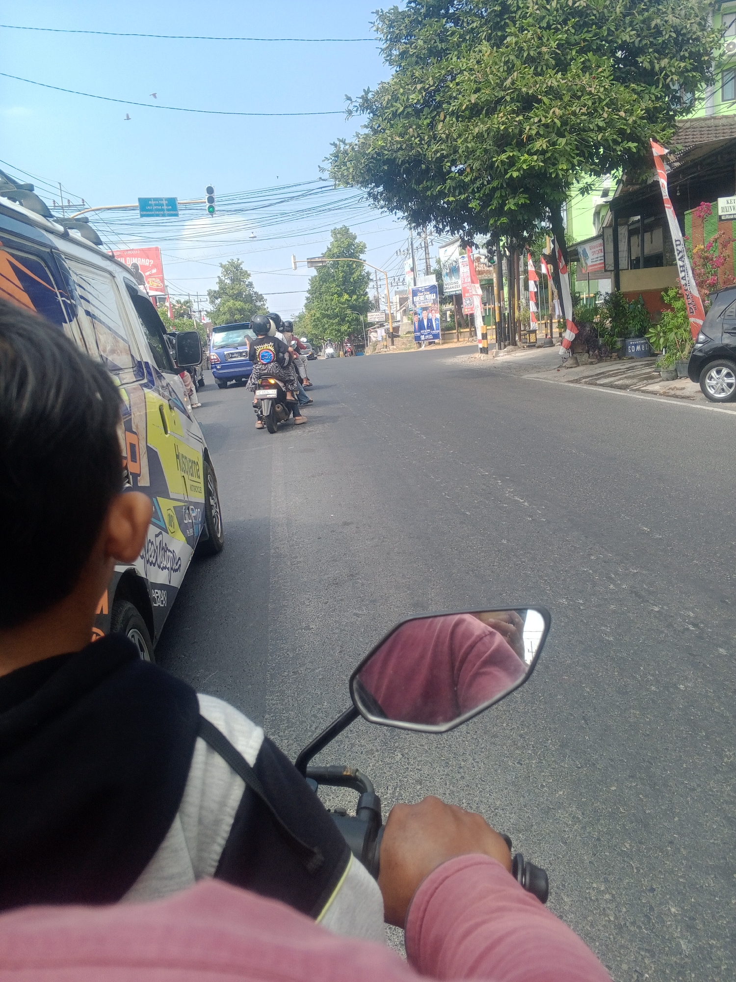 The image captures a street scene in Ronowijayan, Indonesia. A person's shoulder and arm, wearing a pink shirt, are visible in the foreground, holding onto a motorcycle handlebar. A side-view mirror reflects the back of the person's head and shoulders. The motorcycle is in motion on an asphalt road. In the middle ground, several vehicles, including a blue car and a white van with colorful graphics, are on the road. Motorcycles are also present, with riders wearing helmets. The background shows a street lined with buildings, trees, and traffic signals. Indonesian flags and banners are displayed along the roadside. The sky is clear and blue, with a few birds in flight.