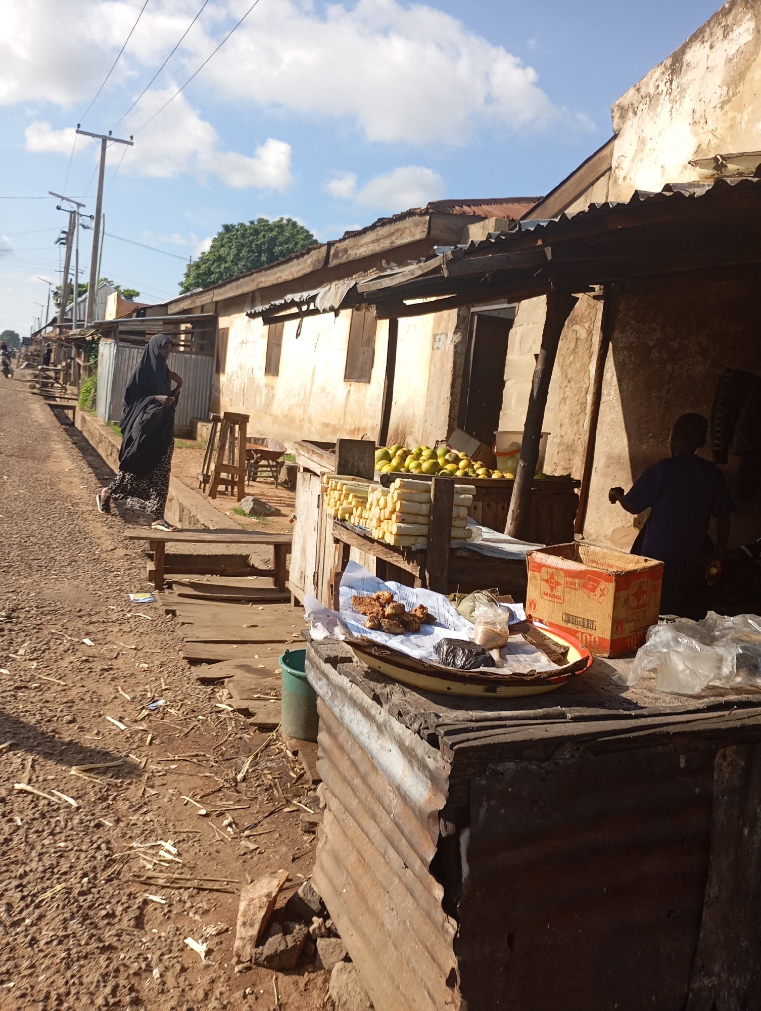 The image depicts a street scene in the city of Shendam, Nigeria, on a sunny day with a partly cloudy sky. A dirt road runs along the left side of the frame, with a woman in a black hijab and patterned skirt walking away from the viewer, towards the left. She is carrying a large black bag over her right shoulder. In the background, several structures with corrugated metal roofs and weathered walls line the road.
To the right of the road, a market stall is set up. Piles of yellow fruits, possibly mangoes or similar, are visible on a wooden table. In front of these, stacks of what appear to be sugarcane stalks are arranged. A shallow dish on a corrugated metal surface contains what looks like fried food items and a dark lump wrapped in plastic. A cardboard box with red and yellow markings, labeled "MAGGI," is also present on this surface, along with plastic bags.
Further to the right, a person in a dark blue shirt is partially visible, facing away from the camera and appearing to interact with the stall. The ground is covered in dirt, small stones, and debris, including what looks like plant matter. A green bucket is visible near the market stall. Power lines are strung across utility poles in the background. The overall impression is of a busy, informal market area.