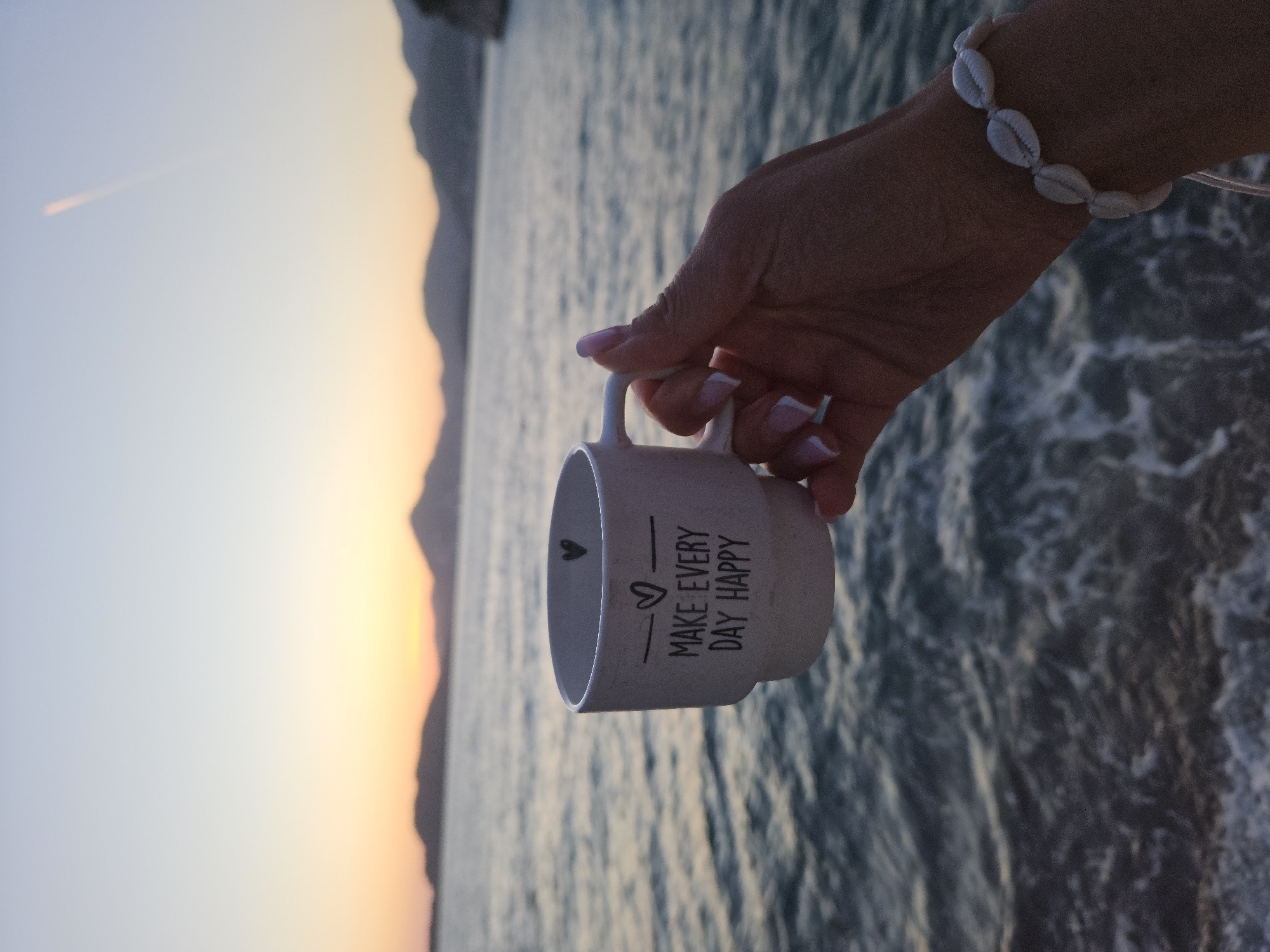 The image captures a serene sunset scene over the sea, featuring a hand holding a mug. The horizon stretches across the image, with calm waters in the foreground and mid-ground gently reflecting the soft evening light. Silhouetted mountains rise along the distant coastline on the left, where the sun is setting, casting warm orange and yellow hues across the lower sky. The sky transitions to a lighter, pale blue towards the upper right. A faint, glowing contrail is visible high in the lighter blue portion of the sky.
In the upper right section of the frame, a person's right hand, with light skin and neatly manicured nails featuring a pink and white French tip design, gently holds a white or light-colored ceramic mug by its handle. A bracelet made of white, shell-like beads adorns the wrist. The mug has black text inscribed on its side, clearly reading "MAKE EVERY DAY HAPPY," accompanied by a small heart symbol.
The scene conveys a peaceful and reflective mood, suggesting the enjoyment of a beverage while watching the sunset over the beautiful coastal landscape of Sveti Stefan, Montenegro. The time of day is clearly sunset, with clear weather.