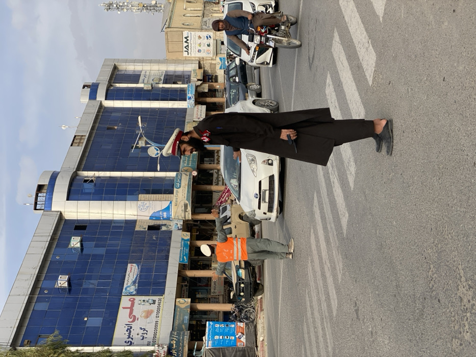 The image depicts an urban intersection in Kandahar, Afghanistan. A paved asphalt road with white zebra-stripe crosswalk markings is visible under a partly cloudy sky.
In the foreground, slightly right of center, a male traffic official stands on the roadway. He wears a dark, possibly black, uniform consisting of a long tunic and trousers, a white peaked cap with a red band, and dark slip-on footwear. He has a dark beard and holds a black rectangular electronic device, possibly a walkie-talkie, in his right hand.
In the midground, positioned centrally within a crosswalk, a second male traffic official is actively directing traffic. He wears light-colored trousers, a light-colored top, a bright orange high-visibility vest, and a white peaked cap. Both of his arms are extended horizontally with palms open, signaling a stop.
Vehicular traffic is present. A white hatchback or small sedan is stopped near the foreground official. Behind the second official, a light-colored pickup truck and another white sedan are visible. On the far right, a man on a silver motorcycle is situated on the road, wearing a dark blue vest over a brown garment.
The background features a multi-story building on the left with a modern facade, characterized by extensive blue reflective glass paneling and distinctive curved architectural elements at its upper corners. Several commercial signs, displaying text in Dari or Pashto script (including "ملی انجنیری گروپ" and "Ariana Cargo") and various images, are affixed to the lower levels. Air conditioning units are mounted on the building's exterior. To the right of this modern structure, older, lighter-colored buildings are visible, and a tall communications tower equipped with multiple antennae stands against the sky on the far right.
