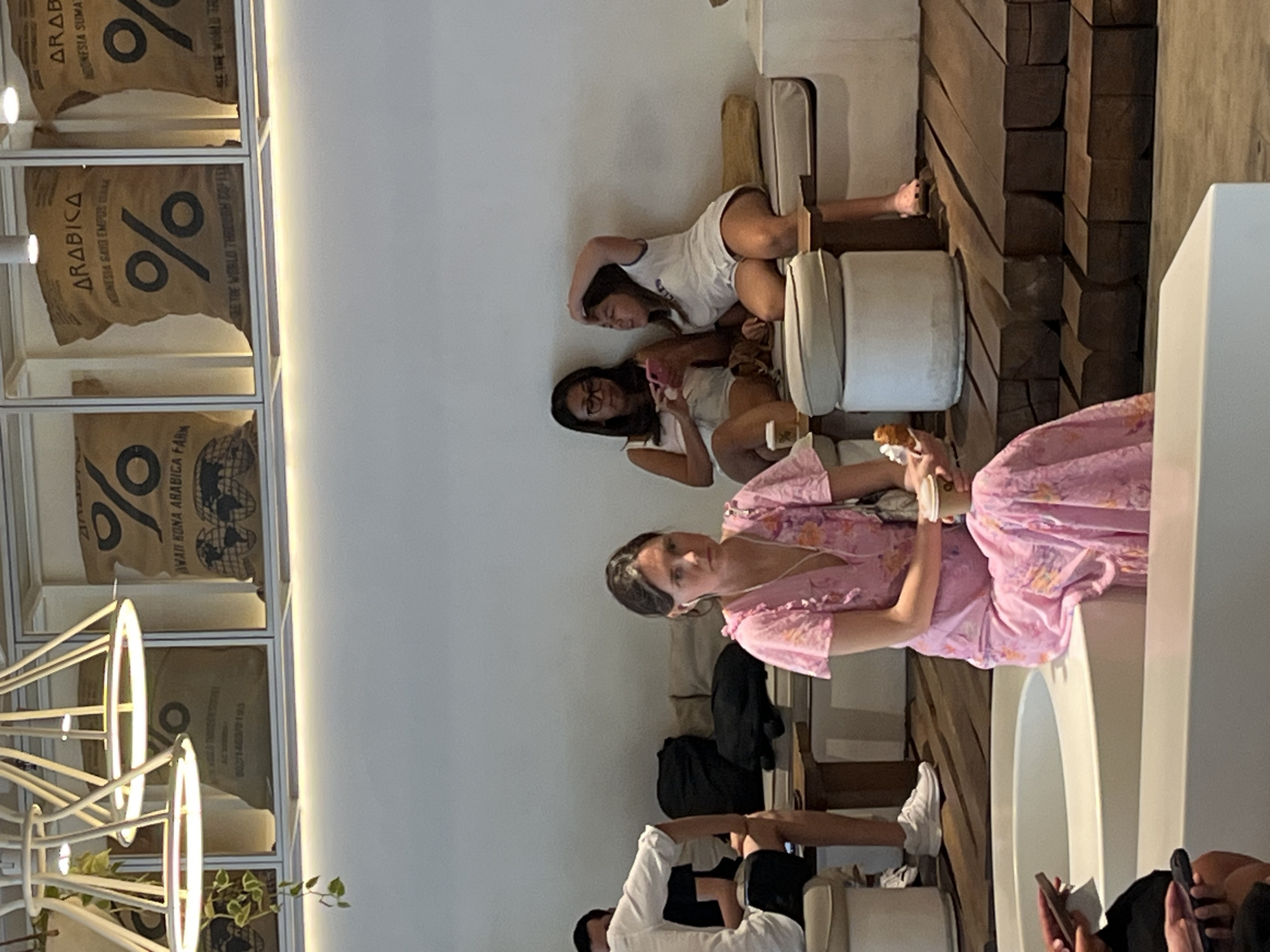 The image shows a group of people inside a brightly lit, modern cafe. In the foreground, a woman in a pink floral dress is seated on a white structure, holding a coffee cup. To her right, a young girl in a white t-shirt and shorts sits with her hand on her head. To her left, another woman wearing glasses and a white top is looking at her phone. Behind them, a man in a white shirt is visible from the side. The cafe has a rustic aesthetic with wooden beams forming a tiered seating area and large burlap bags with "ARABICA" and "%" logos displayed on the wall. The overall impression is that of a relaxed social gathering in a stylish cafe, likely located in Kuta, Indonesia, given the context.