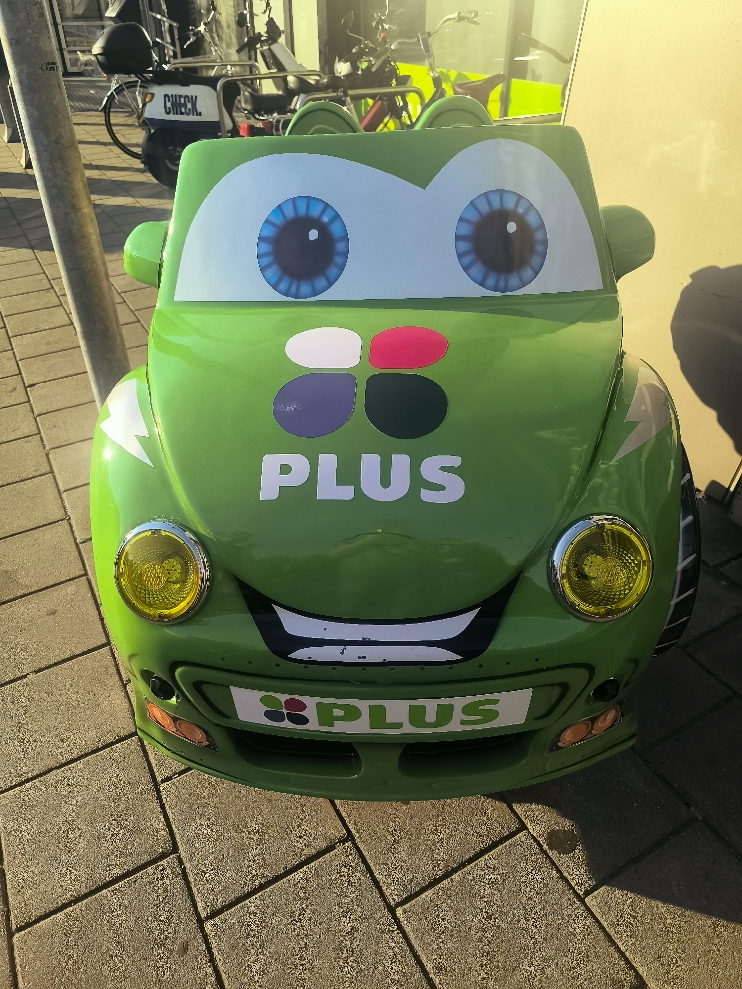 The image shows a brightly colored, cartoon-style green car-shaped play structure situated outdoors on a paved surface. The play structure has large, expressive blue eyes and a wide smile. It is adorned with colorful polka-dot graphics and white text that reads "PLUS" on its hood and a similar graphic with "PLUS" on a white license plate below. The car has yellow headlights and orange indicator lights. In the background, there are bicycles and a scooter parked, with a building visible behind them. The setting appears to be a public area in Almere, Netherlands, possibly near a residential or commercial zone, indicated by the presence of the parked vehicles and the urban paving. The sunlight suggests it is daytime.
