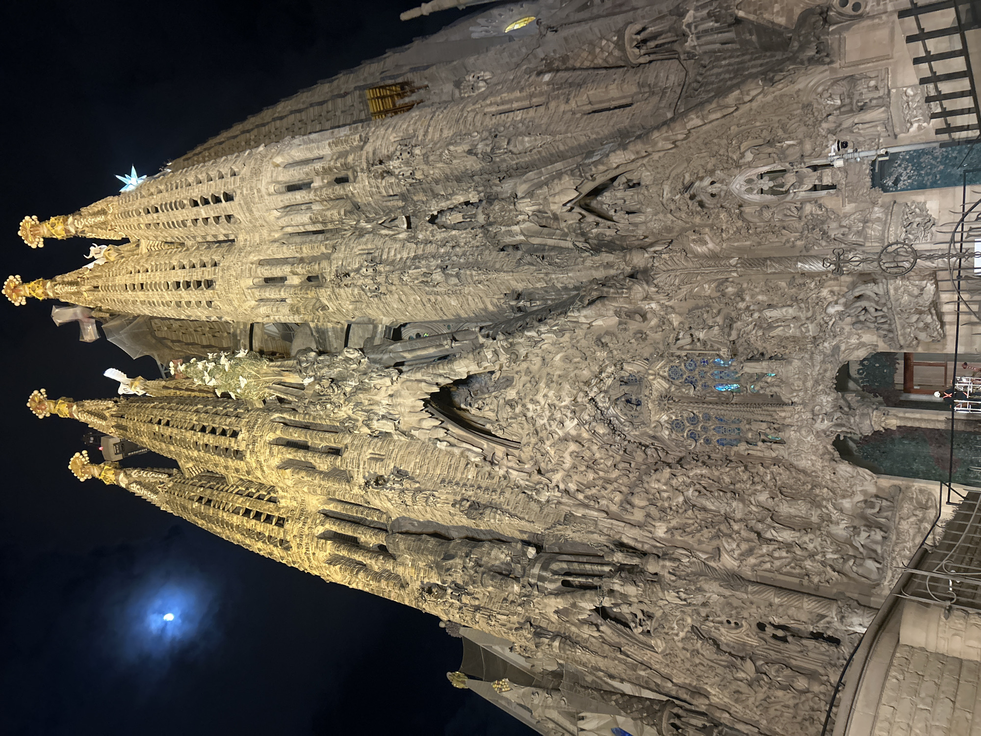 The image captures the Sagrada Familia basilica in Barcelona, Spain at night. The structure’s intricate facade is illuminated, showing the detailed stonework and sculptures. The cathedral's towers are visible, with one crowned with a large, illuminated star. A faint moon appears in the dark sky in the bottom left corner of the frame. The foreground includes architectural elements and what appear to be parts of an entrance or boundary, like ironwork. No people are visible in the frame.