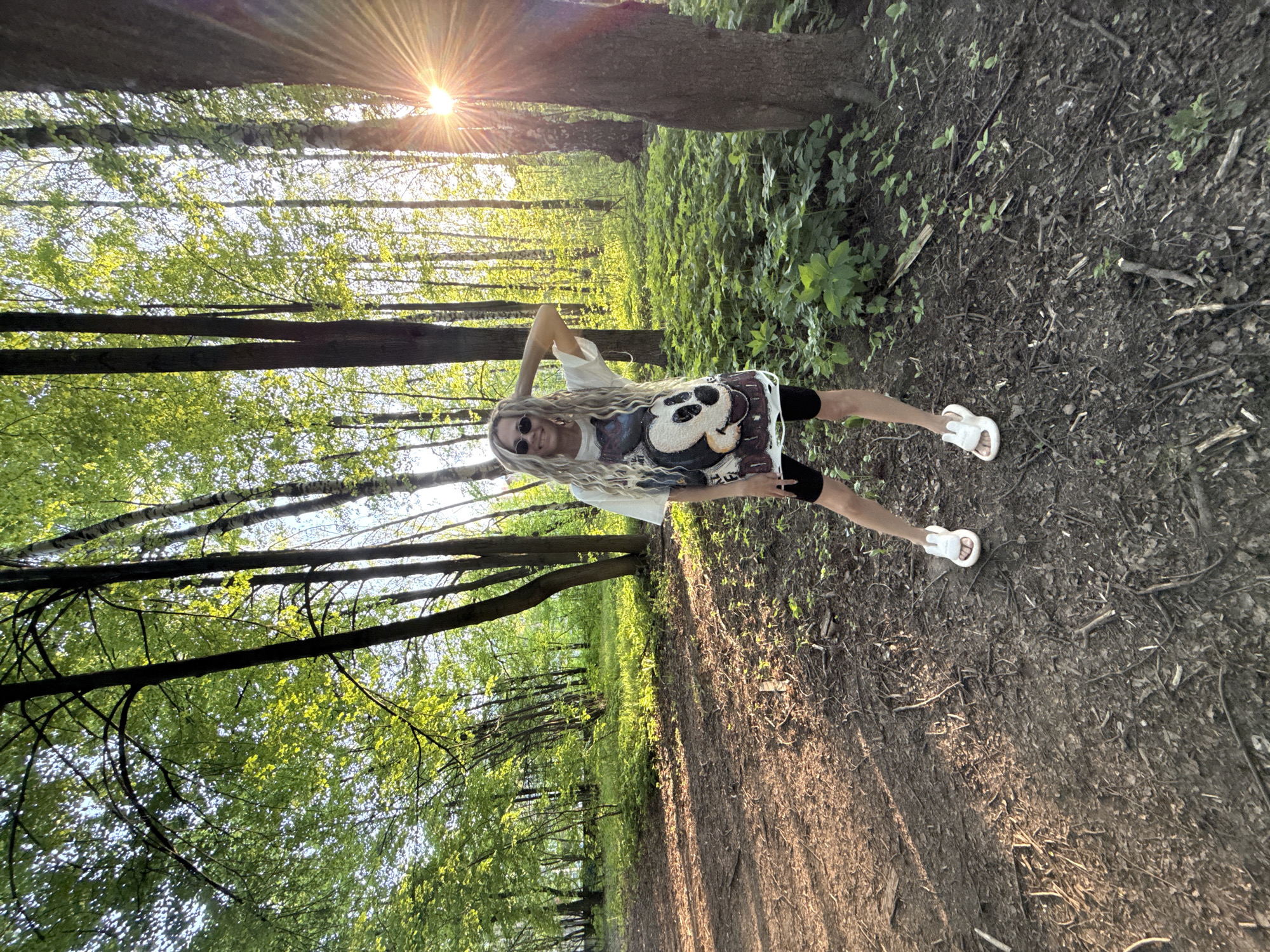 The photo shows a woman standing in a wooded area, likely a park, in Шебунино, Russia. She has long, blond hair, wears sunglasses, a t-shirt with a graphic design, black shorts, and white sandals. She is posing for the photo with one hand raised behind her head. The area is a forest with sunlight streaming through the trees. The ground is covered in dirt, leaves, and some vegetation. The sun is visible through the trees in the background.