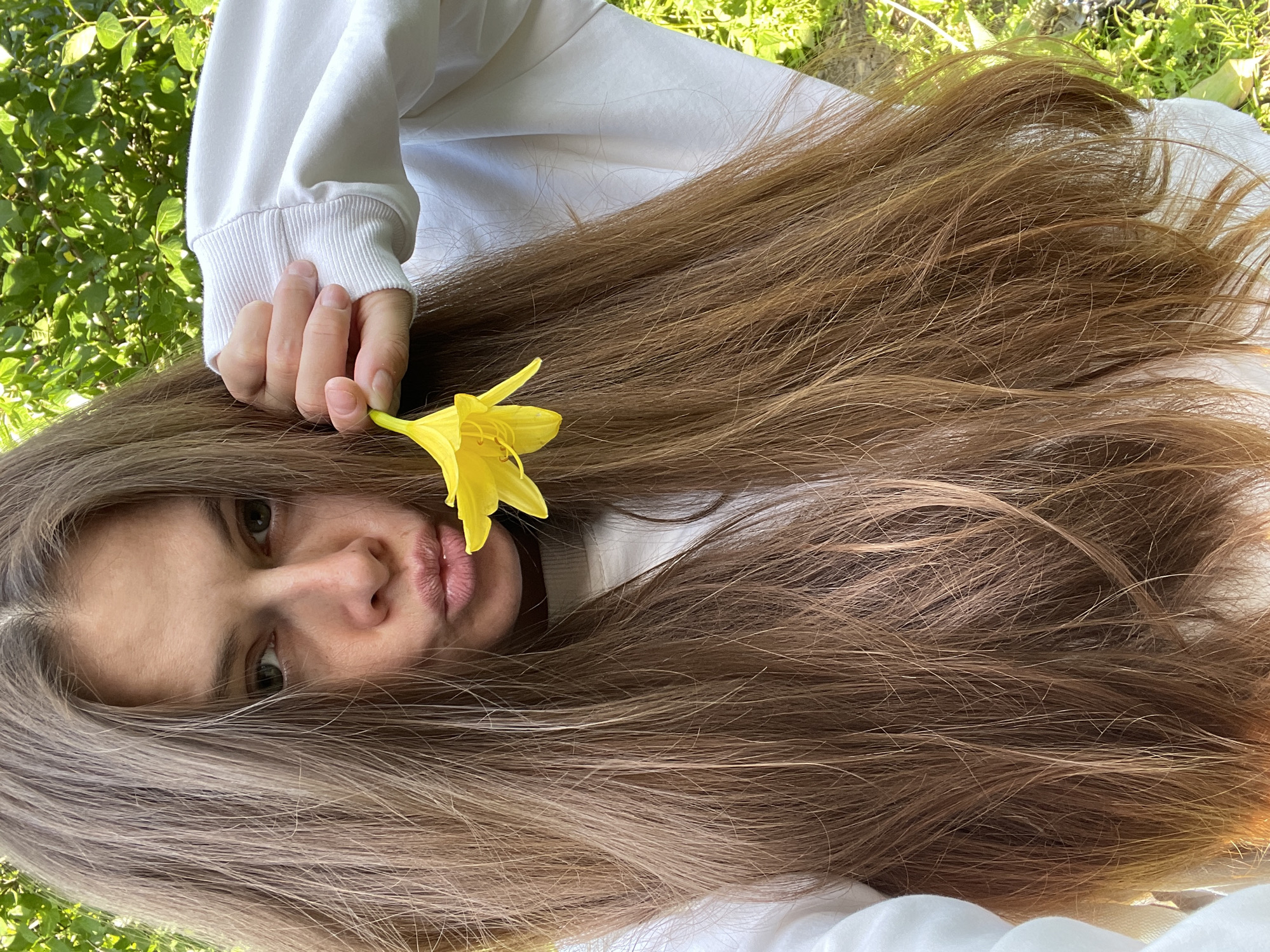 A young woman with long, brown hair is taking a selfie outdoors. She is wearing a white sweatshirt and holding a yellow flower near her face, with her lips pursed. The background consists of green foliage, suggesting a natural or garden setting. The lighting is bright, indicating it is daytime. The location is not specified to be in the city of Заречный, Russia, as there are no identifying features in the image to support this claim.