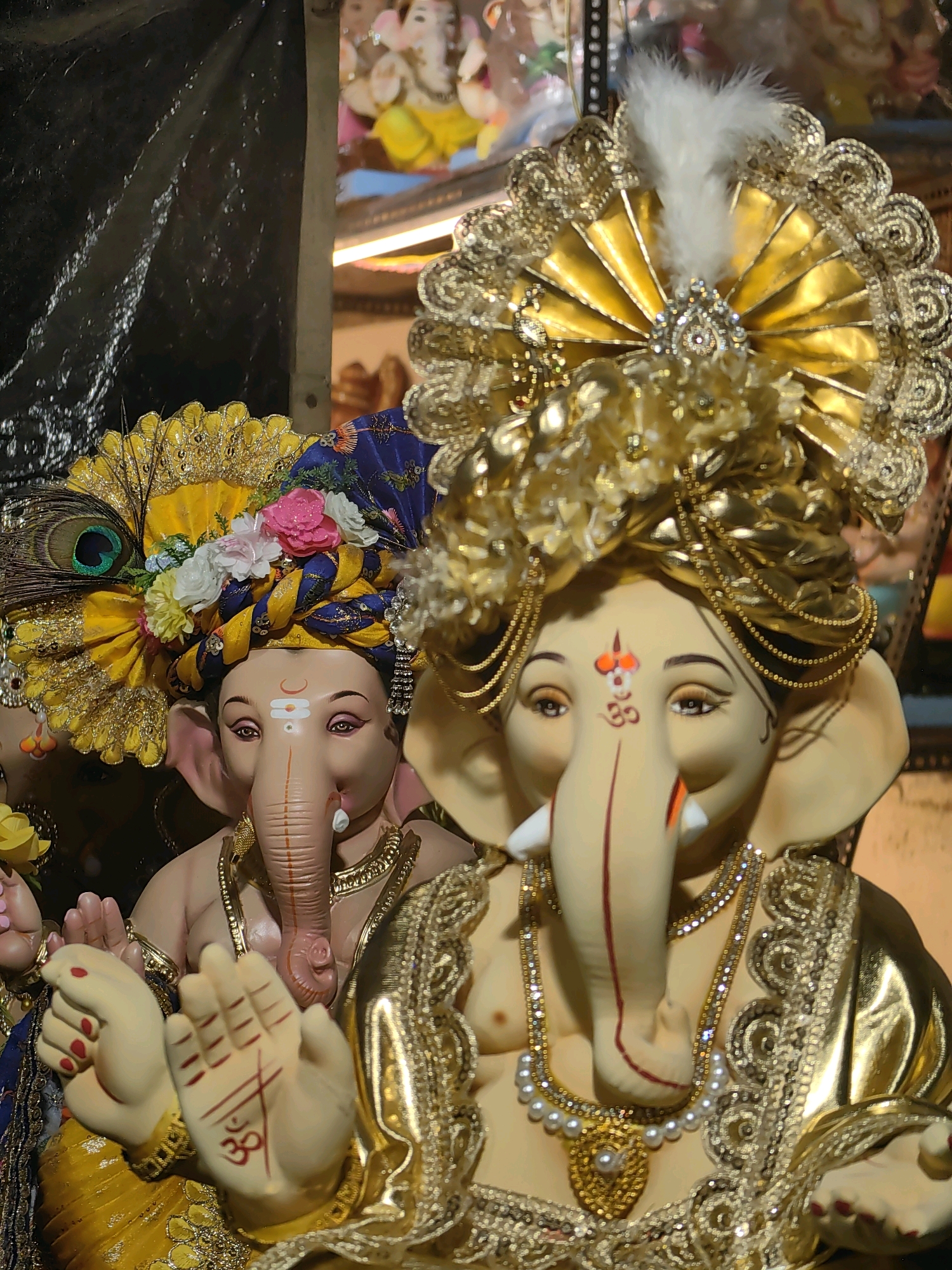 Two clay idols of the Hindu deity Ganesha are displayed prominently in the foreground, both adorned with elaborate golden headdresses and ornate jewelry. The idol on the right, closer to the viewer, is partially obscured by its headwear, featuring a white feather plume and intricate gold lacework. It wears a pearl necklace and a golden garment embellished with sequins and lace. The idol on the left, further back and slightly to the left, is also decorated with a vibrant turban featuring floral and peacock feather accents. Both idols have their hands raised, with the idol on the right displaying an 'Om' symbol and lines drawn on its palm. The background is dark and out of focus, with hints of other decorations and possibly another Ganesha idol visible. The scene appears to be from a festival or religious gathering, likely during Ganesh Chaturthi celebrations in Mumbai, India, given the cultural significance of the deity and the common practice of displaying such idols during this time.