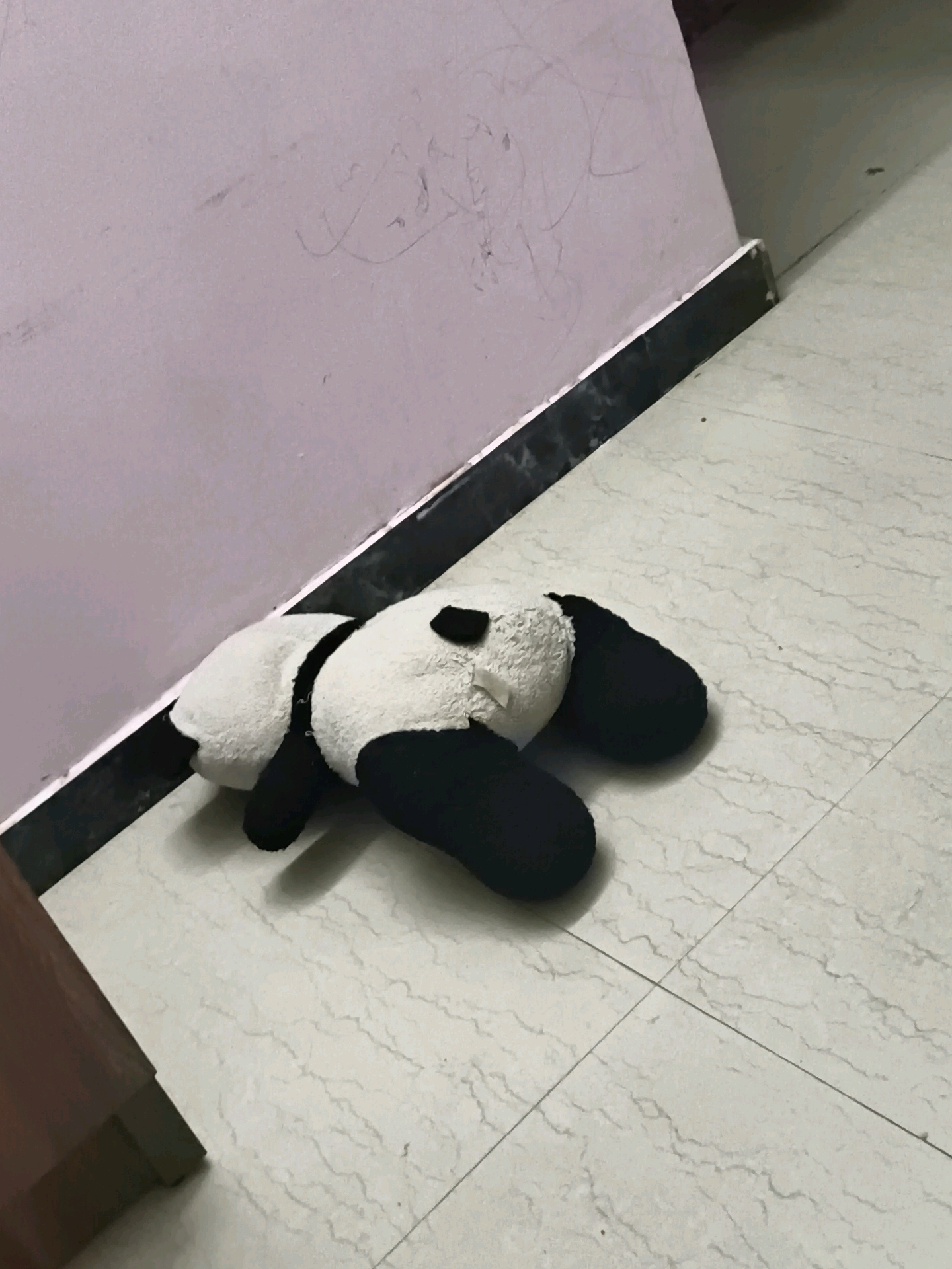 A plush panda toy lies on its stomach on a tiled floor next to a wall with a black baseboard. The panda is black and white, with black patches around its eyes and ears, and black limbs. The floor tiles are light-colored with a marble-like pattern. The wall is a light purple color and has some faint, scribbled markings on it. The scene appears to be indoors. There is no indication of the city of Coimbatore, India.