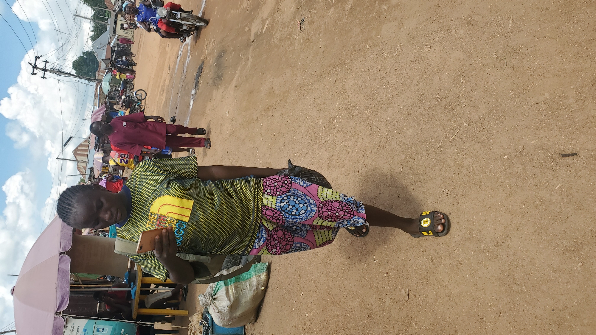 A young Black woman is walking on a dirt road in Jalingo, Nigeria, looking at her cell phone. She has braided hair and is wearing a yellow and green t-shirt with a yellow design on it, and a patterned skirt. She also has a bag on her shoulder and is carrying a black plastic bag in her right hand. In the background, there are several market stalls with people walking and riding motorcycles. The sky is cloudy.