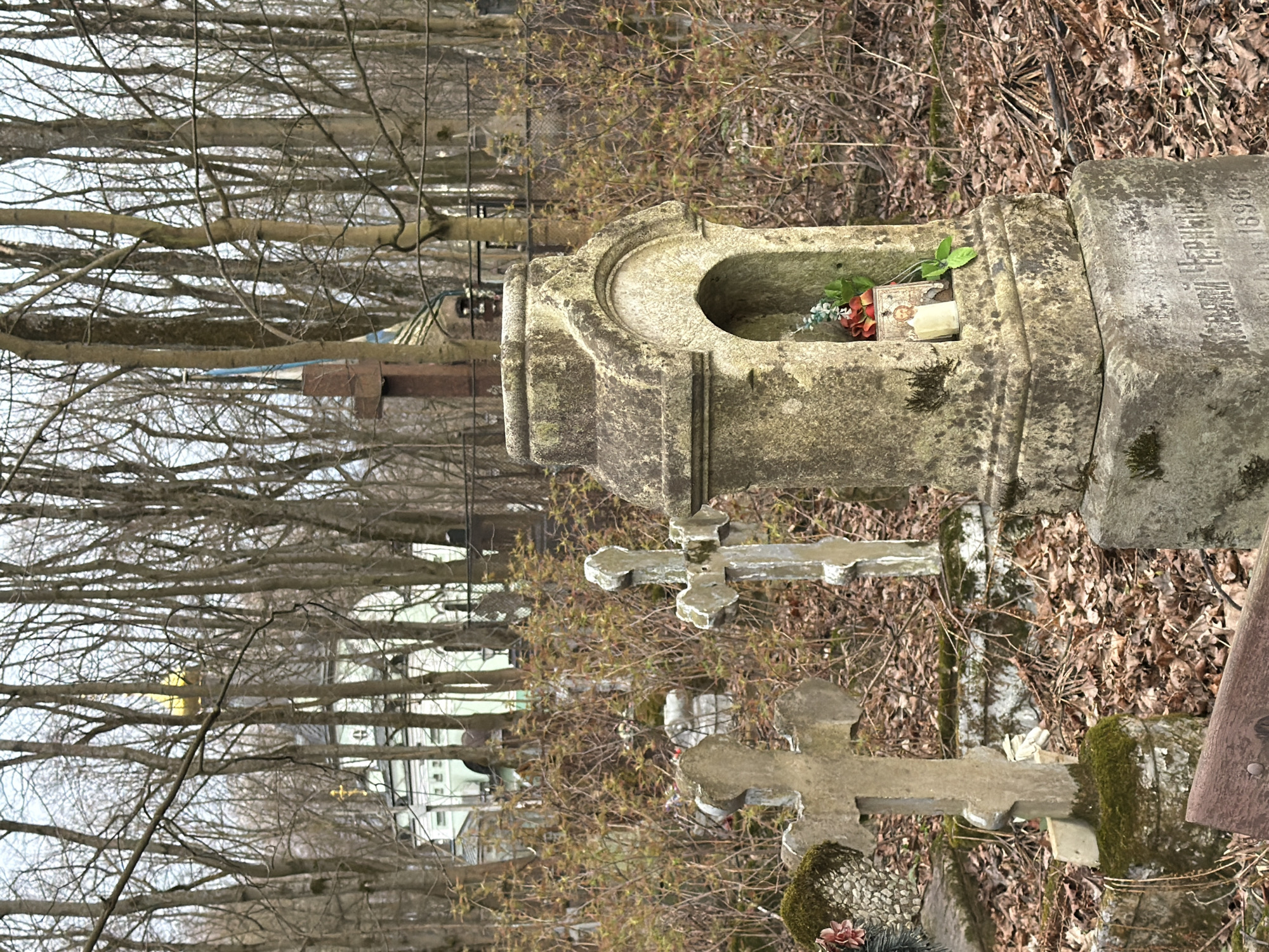 The image shows a graveyard in what appears to be Saint Petersburg, Russia, based on the Cyrillic script on the gravestones. The foreground features weathered stone monuments, including a niche-like structure holding artificial flowers and a religious icon. Several stone crosses stand among the graves. Leaf litter covers the ground. In the background, bare trees and buildings are visible. The light suggests it is either early morning or late afternoon. There are no people present in the frame.
