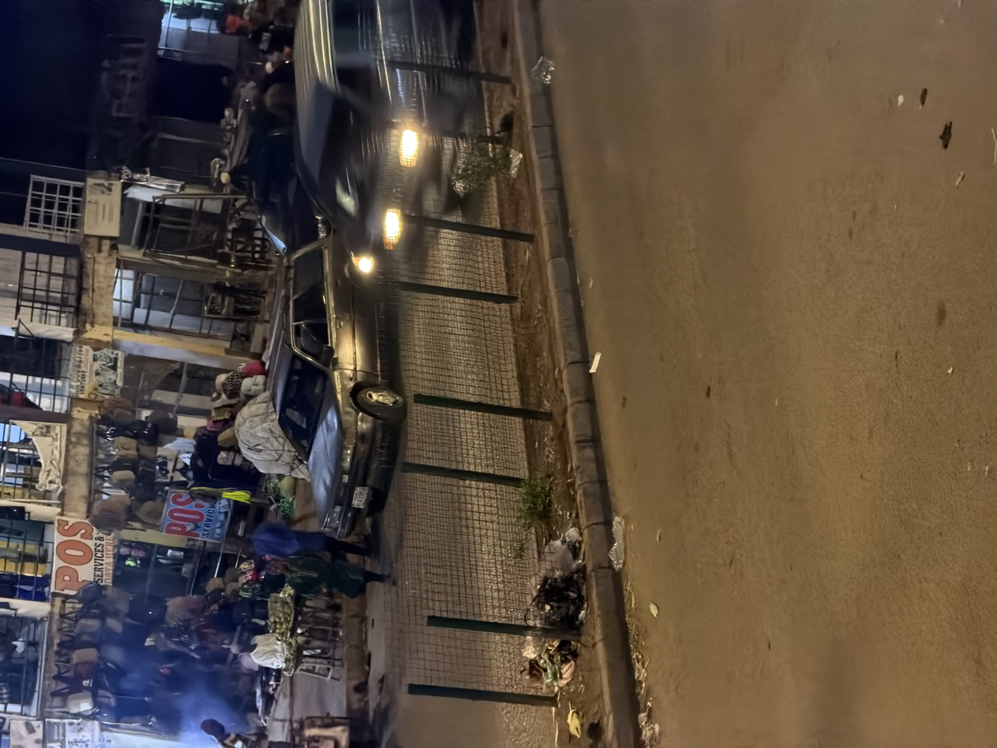 In Bukuru, Nigeria, a busy street scene unfolds at night, illuminated by streetlights and the glow of businesses. A gray sedan is parked at an angle in the foreground, its headlights casting a warm glow. A dark-colored van, also with headlights on, is in motion behind it, its form blurred by movement. The street is lined with businesses, some featuring bright "POS" signs indicating point-of-sale services. Bags and merchandise are displayed outside these establishments, suggesting a commercial area. Pedestrians are visible, including a person in a green garment walking near a stall laden with produce. A wire fence separates the street from a strip of land where some trash has accumulated, including plastic bags and debris. The overall impression is one of a lively, bustling urban environment at dusk.