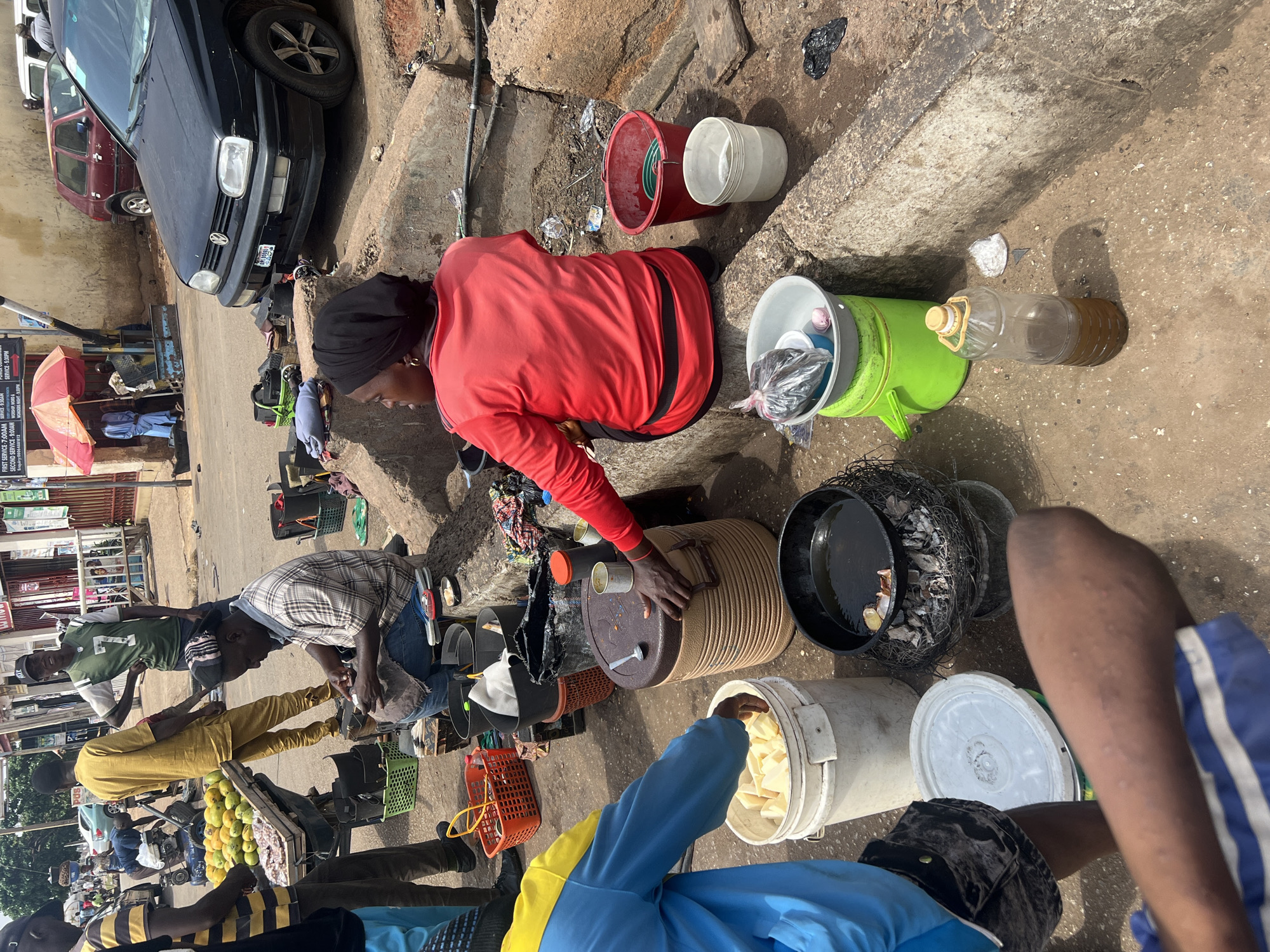 This image depicts a bustling outdoor street market scene in Gura Top, Nigeria, captured under bright daylight. The ground is a mix of dirt and pavement with concrete curbs, suggesting an informal commercial area.
Several individuals are engaged in various activities. A prominent woman in a red long-sleeved shirt and black head covering is seated on the ground or a low stool, actively tending to several containers. These include a large, ribbed brown storage bin, a black pot with charcoal beneath it (indicating a cooking or heating setup), and various plastic buckets. One green bucket contains packaged items, and a nearby plastic bottle holds a dark liquid, possibly cooking oil. In the immediate foreground, a person wearing a blue and yellow top reaches into a white bucket filled with cut white pieces, likely a food item such as yam or cassava. Further back, a man in a plaid shirt and jeans kneels or bends over, handling goods, while other people mill about, some near stalls or vehicles.
Objects visible include a black Volkswagen sedan and a red car parked along the side, an orange umbrella providing shade for a stall, various baskets (some containing green-yellow fruits, possibly mangoes), and general market debris scattered on the ground.
A prominent, rotated sign in the background lists church service times: "FIRST SERVICE-7:00AM," "SECOND SERVICE-9:00AM," "SERVICE-5:30PM," and "WORSHIP WORD A." It also displays "WOADERS NIGHT-9:30PM" and an "Enquiry" number: "00064881812." The overall scene captures the vibrant, everyday commerce and social interaction typical of a West African market.