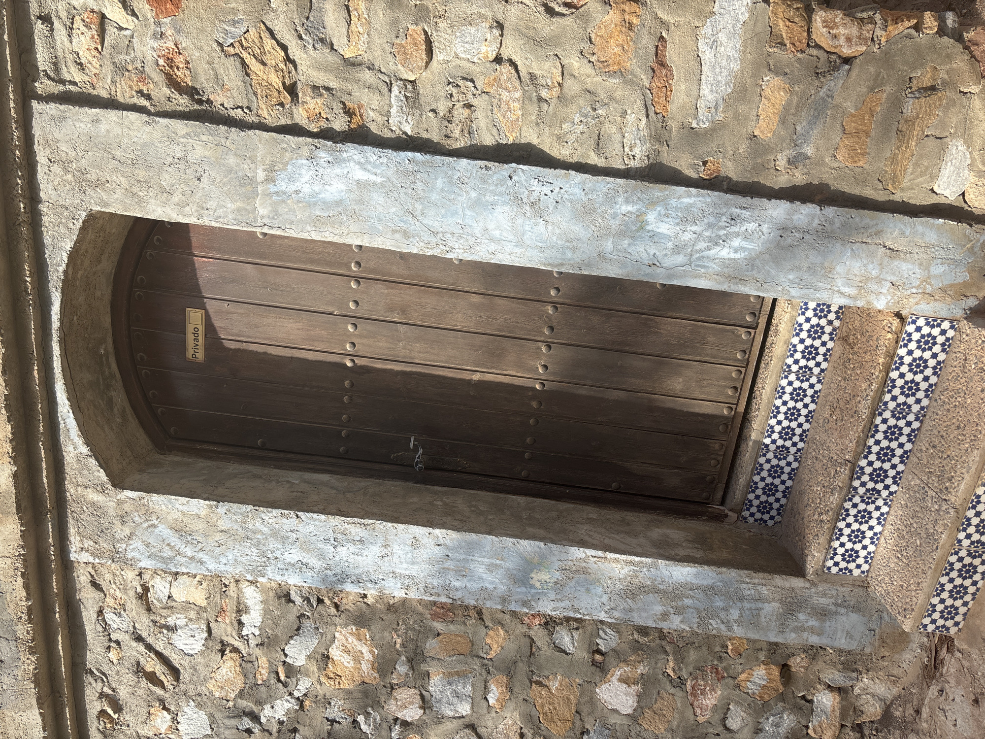 An arched wooden door, labeled "Privado," is set into a rough stone wall. The door is framed by a textured, slightly worn plaster archway. A small, dark metal latch is visible on the door. In front of the door, a short set of stairs ascends, with each step edged in decorative blue and white tiles featuring a starburst pattern. The surrounding wall is constructed of irregular stones of varying sizes and colors, mortared together with a rough, uneven grey cement. The lighting suggests it is daytime, with strong sunlight casting shadows on the scene. The environment appears to be outdoors, possibly in a Mediterranean or similar climate, given the stone and tile work.