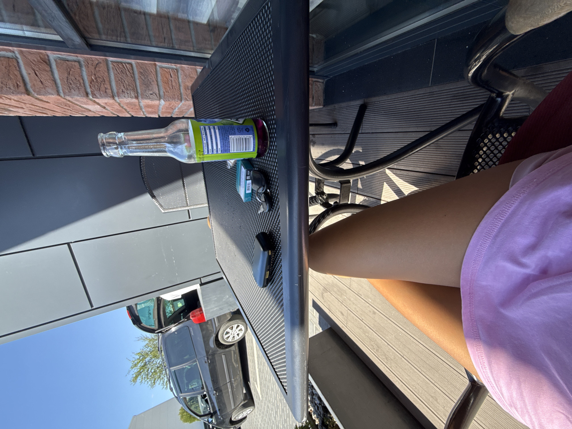 The image depicts an outdoor seating area, possibly a cafe or restaurant patio, with a person's legs and lower torso visible in the foreground, seated in a chair. They are wearing a pink garment, possibly shorts or a skirt. A dark-colored table with a textured surface is in front of the chair. On the table are several objects: a glass bottle with a green label containing a purple liquid, a cigarette pack, a small black electronic device, and a set of keys. In the background, a dark-colored minivan with its rear hatch open is parked. The overall environment suggests a sunny day, with shadows cast by the table and the building. The building's facade features dark gray paneling and red brickwork. The ground beneath the table and chair is a wooden deck. The location is identified as Comrat, Moldova.