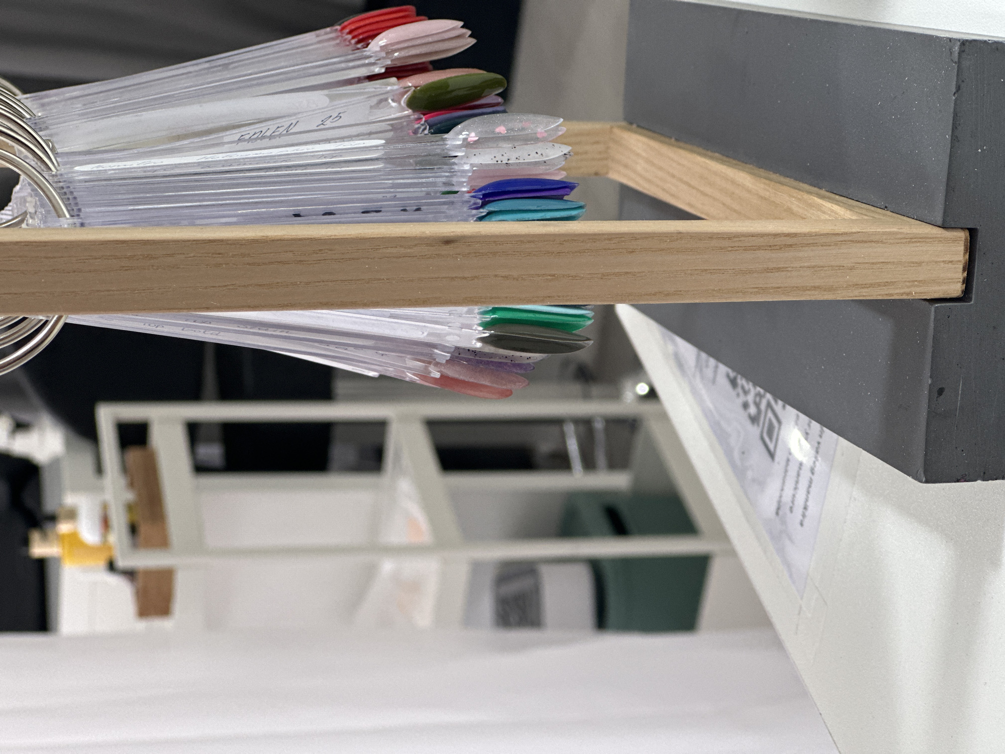 The image, rotated 90 degrees counter-clockwise, captures a close-up of various nail polish color samples displayed on clear plastic swatches. These swatches are organized on multiple metal rings, hanging from a light-colored wooden bar. The wooden bar is securely fitted into a groove within a dark gray, rectangular display block, suggesting a sturdy and professional setup.
A diverse range of nail polish colors and finishes are visible, including shades of pink, red, olive green, light blue, teal, dark blue, dark green, and variations with glitter (silver with heart confetti, clear with black specks). Many of the clear plastic sticks have handwritten labels; one prominent label reads "EPIEN 25." Faint text like "top coat" is also visible on other sticks.
Below the dark gray block, a white surface is visible, likely a counter or shelf. A piece of paper with a QR code and some indistinguishable text in Latin script is taped to this white surface. The background is blurred, showing additional white shelving units and other items, indicating an indoor commercial setting such as a nail salon or beauty supply store in Budva, Montenegro, where customers would select desired colors. No people or active events are depicted, only the static display of products.