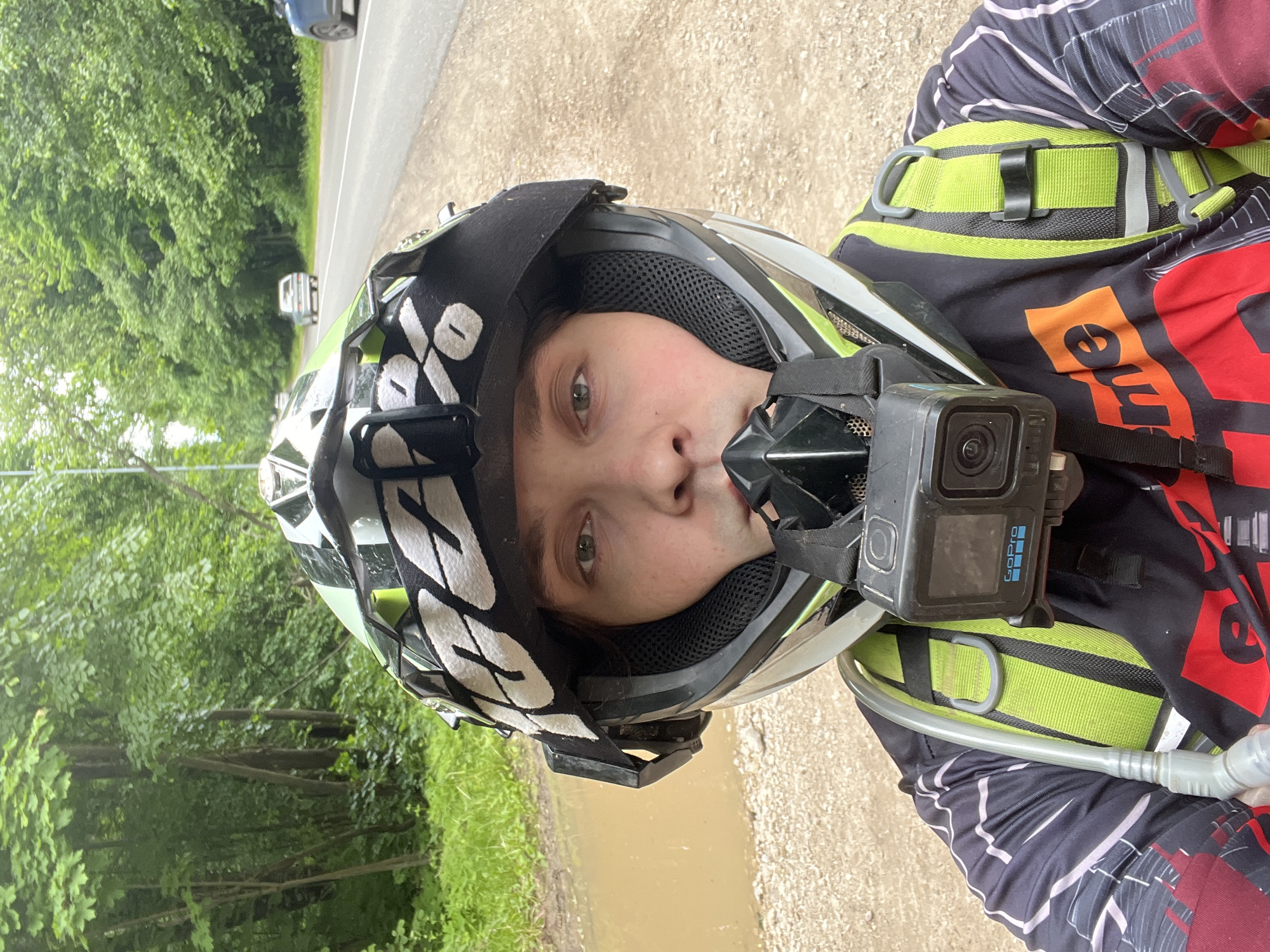 A person is depicted in a close-up selfie-style photograph, standing on a dirt and gravel path adjacent to an asphalt road in Ореховка, Russia. The individual is wearing a full-face off-road helmet with a green, white, and black design, featuring a "100%" brand goggle strap across the forehead area. A black GoPro camera is mounted to the chin bar of the helmet. The person's eyes are visible through the helmet's eye port, while the lower face is obscured by the chin guard and camera mount. Below the helmet, a long-sleeved athletic jersey with a black, red, and orange pattern (partially showing "me" from a word like "extreme") is worn. Over the jersey, a neon green and black hydration pack harness is visible, with a clear hydration tube and bite valve positioned on the left side of the frame.
The environment to the left of the subject features dense green foliage and trees, characteristic of a forest, along with a muddy, light brown puddle on the ground. To the right, an asphalt road stretches into the distance, bordered by more trees, and shows several vehicles (two light-colored cars, one blue car) in motion. The sky is overcast, and the overall scene suggests a daytime setting.