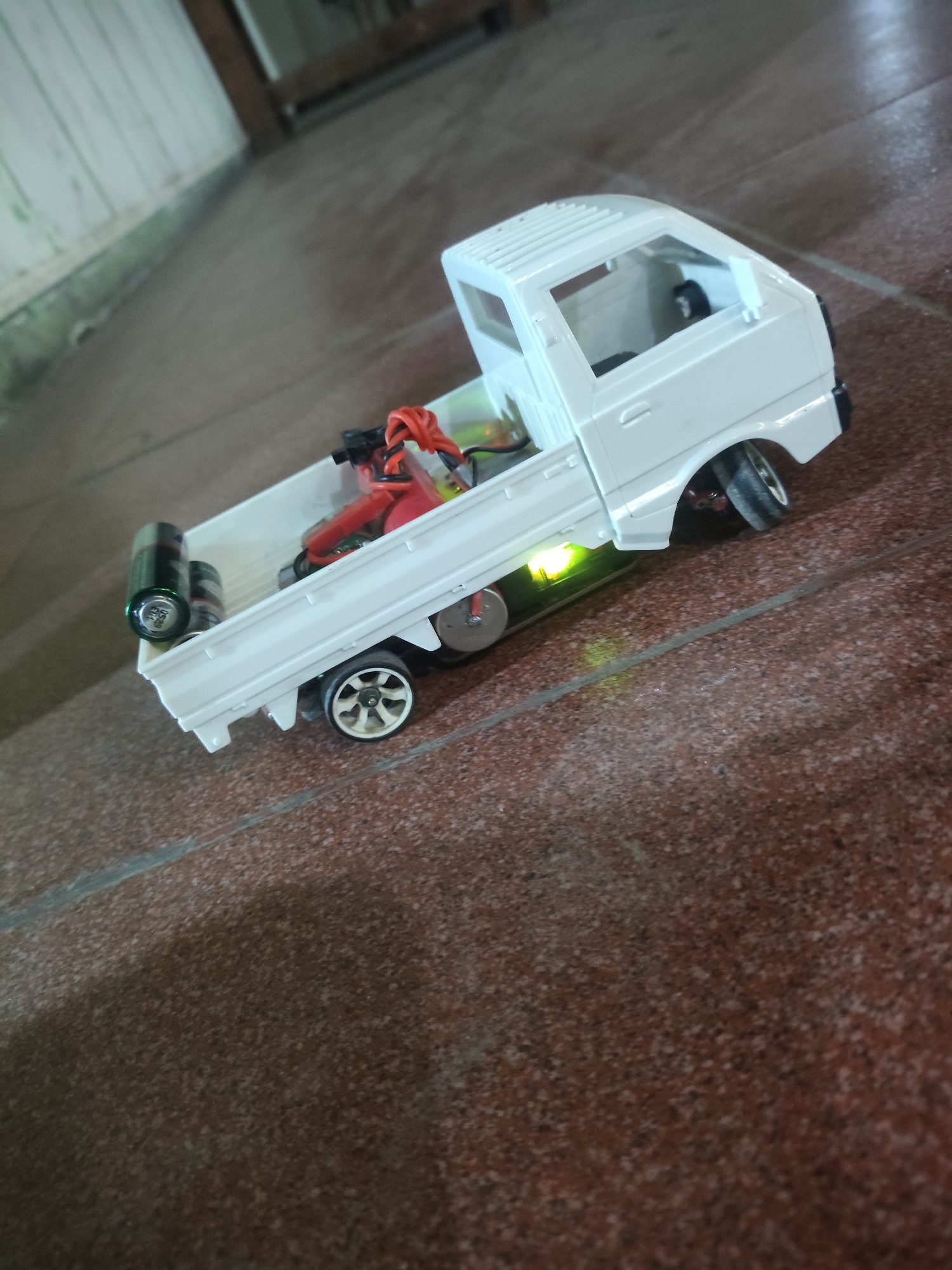 A close-up, high-angle shot shows a white toy truck with an open bed sitting on a tiled floor. Two AA batteries and some red and black wires are visible in the truck bed. A small green light is illuminated on the underside of the truck. The truck has silver wheels with black tires. The floor is tiled with reddish-brown tiles and grout lines. In the background, a wooden surface and a white wall are visible. The image appears to have been taken indoors, possibly in a residential setting in Sei Mencirim, Indonesia, based on the visual cues.