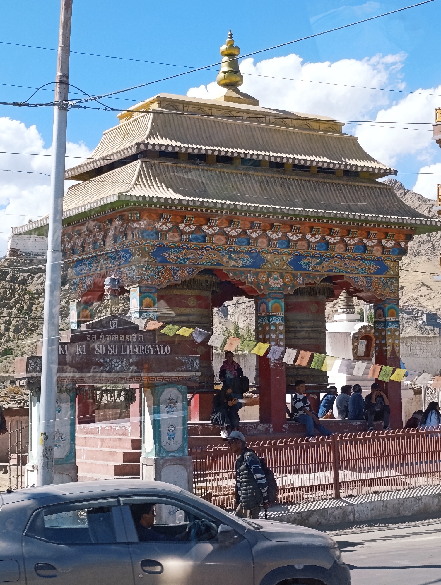This is a street-level, eye-level, medium-shot photograph of a prayer wheel structure in Leh, India. The structure features a golden finial and intricate painted designs on its exterior, with prayer flags strung across the front. A sign in both Tibetan and English reads "KI KI SO SO LHARGYALO". Several people are gathered around the structure, some seated on a bench behind a red metal fence, while others stand. In the foreground, a dark gray car is partially visible, with a person in the driver's seat. The background shows a rocky hillside with some buildings and clear blue sky with scattered clouds. The overall scene suggests a public space in Leh, likely a place of cultural or religious significance.
